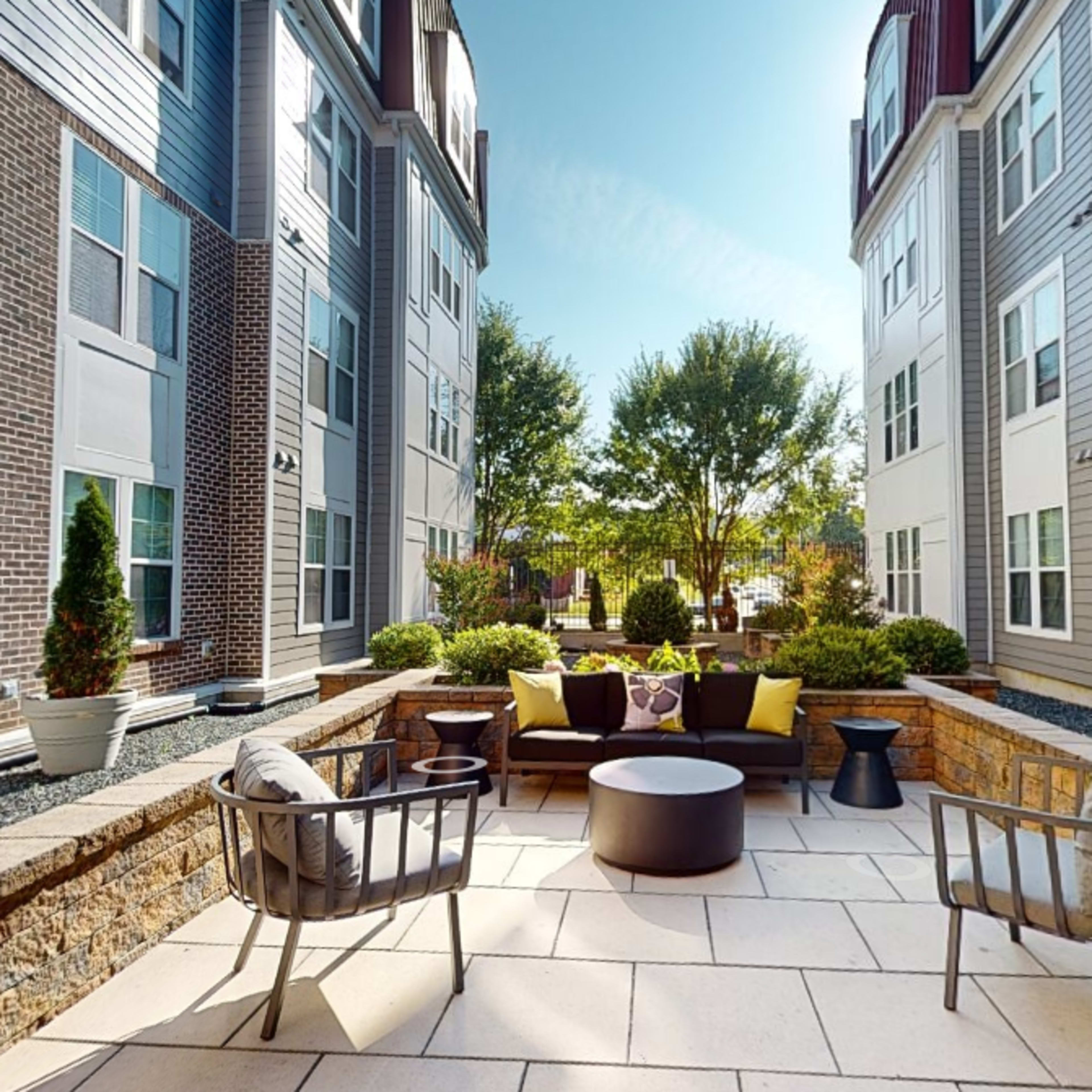 A paved outdoor patio area features seating arrangements and landscaped greenery between two modern apartment buildings.