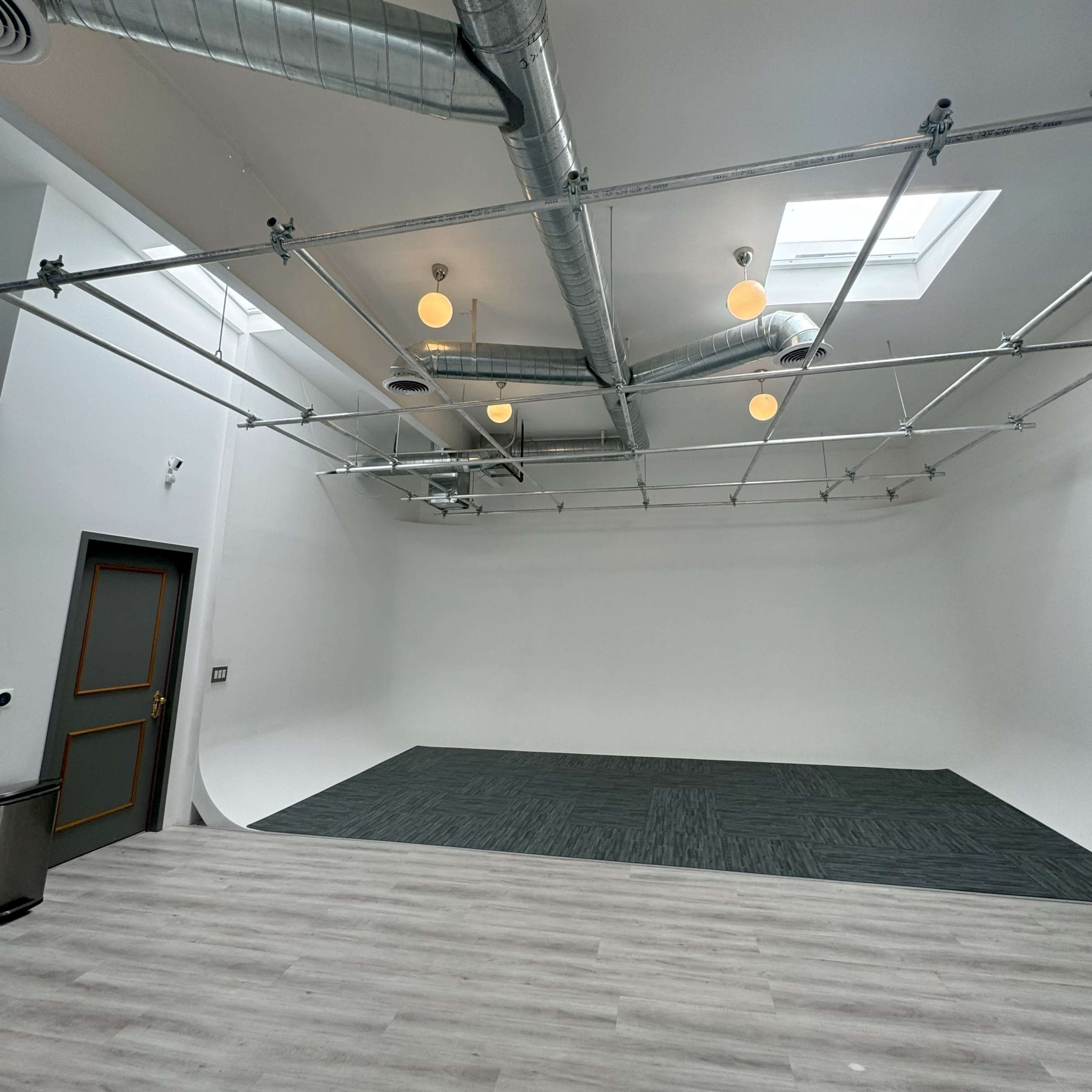 A spacious, empty studio with a white backdrop, gray flooring, and visible ceiling ducts and lights.