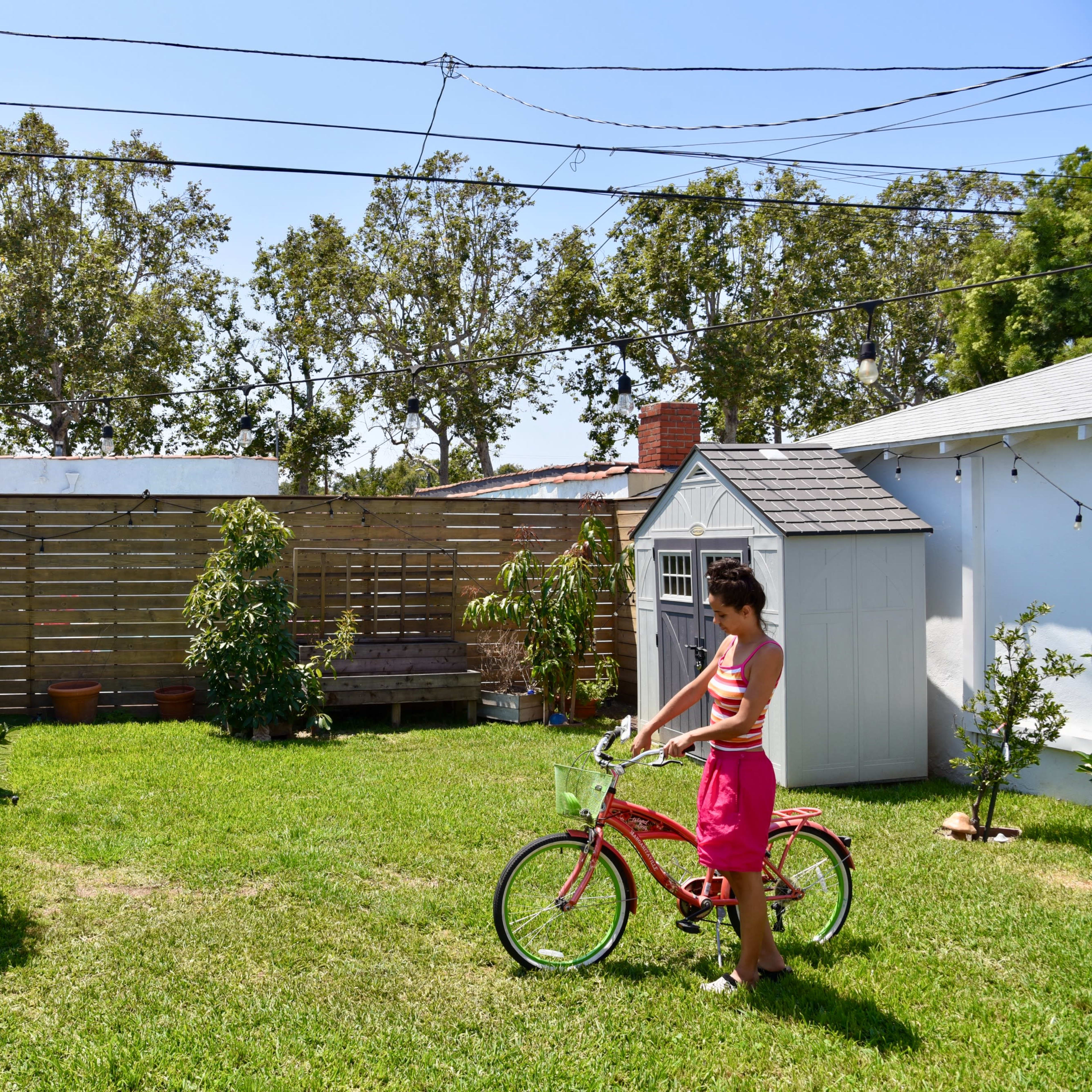 A child stands beside a pink bicycle in a grassy backyard surrounded by small structures and trees.