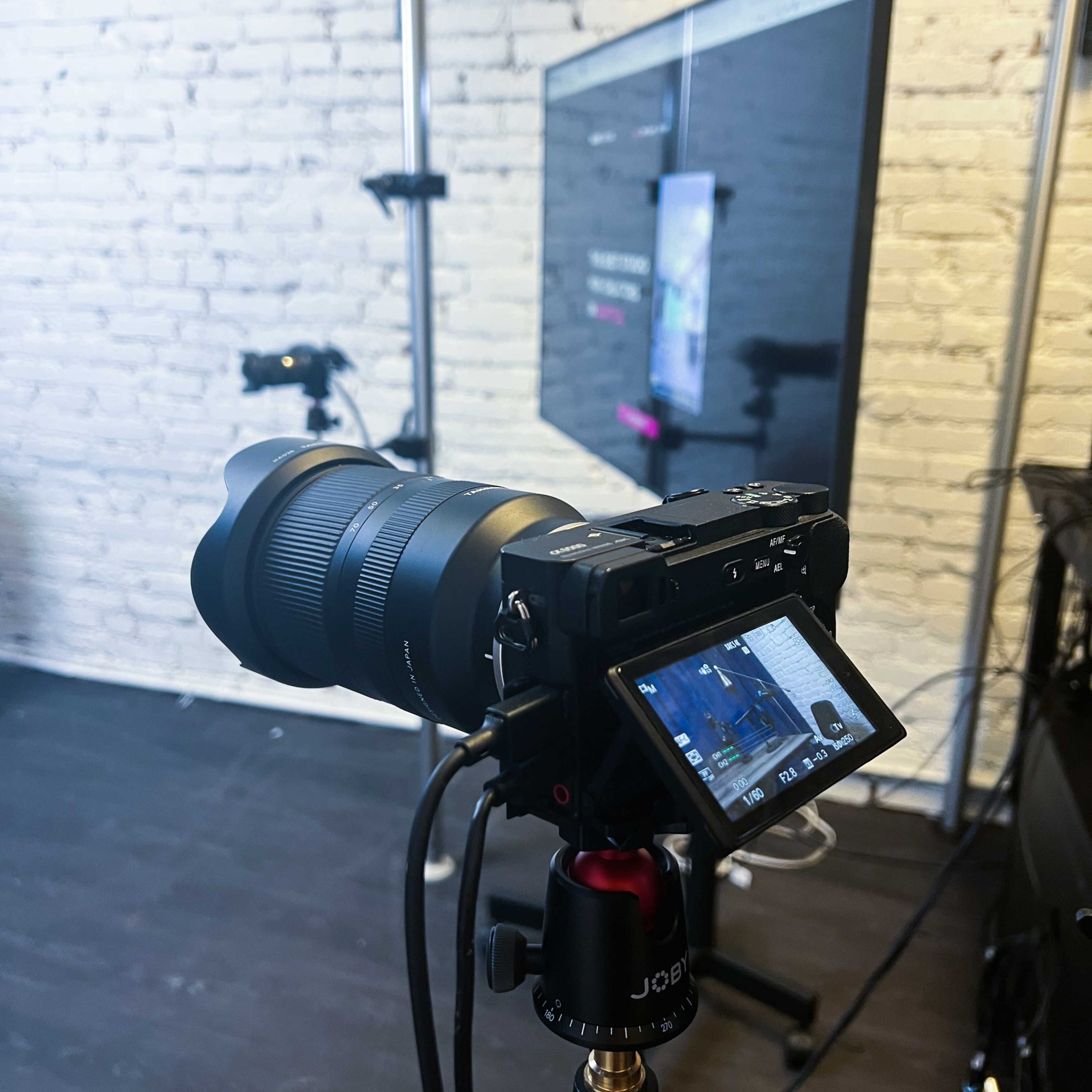 A camera is positioned on a tripod, focused on a monitor displaying an interface, with white brick walls in the background.