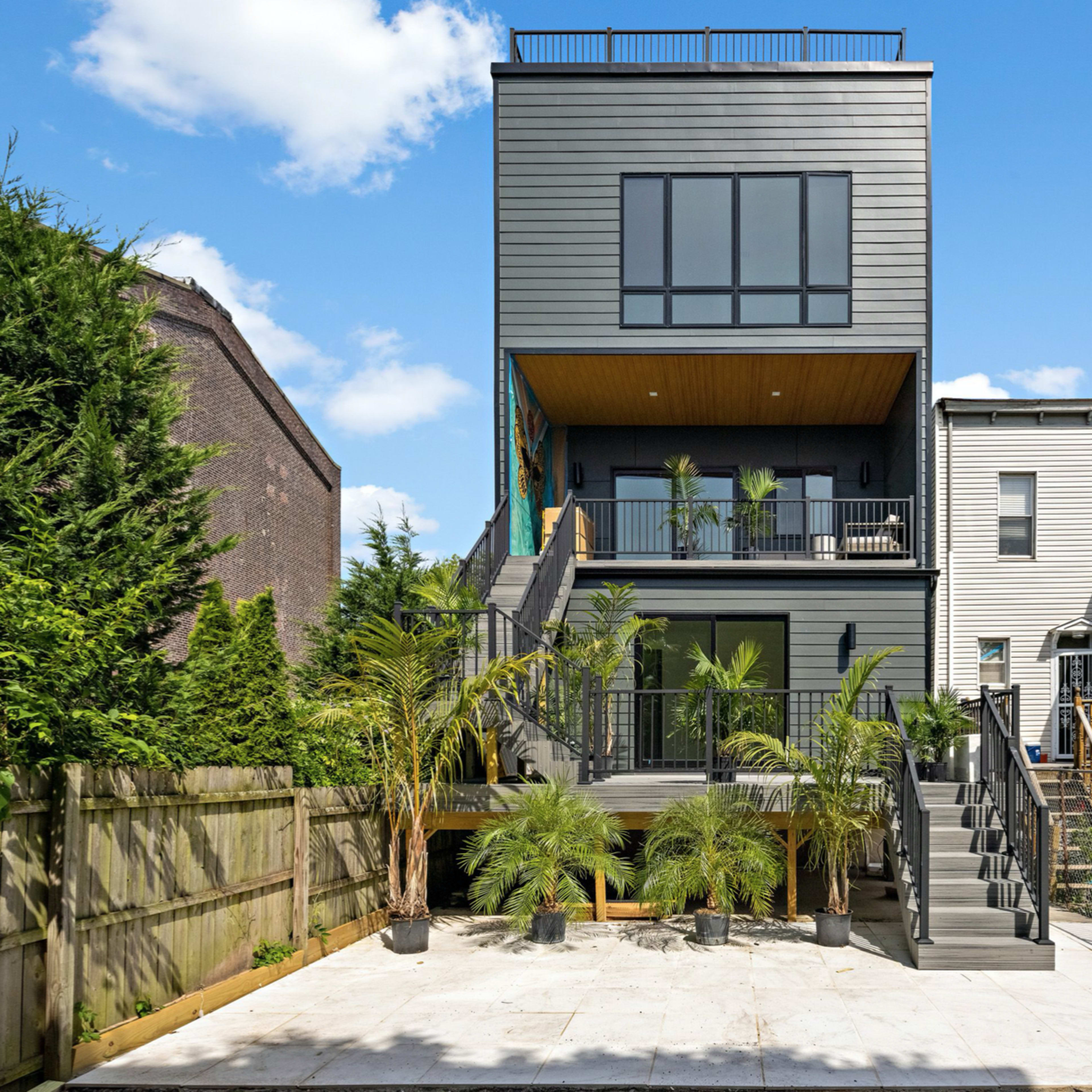 A modern three-story house with a rooftop deck, surrounded by greenery and featuring a paved outdoor area with potted plants.