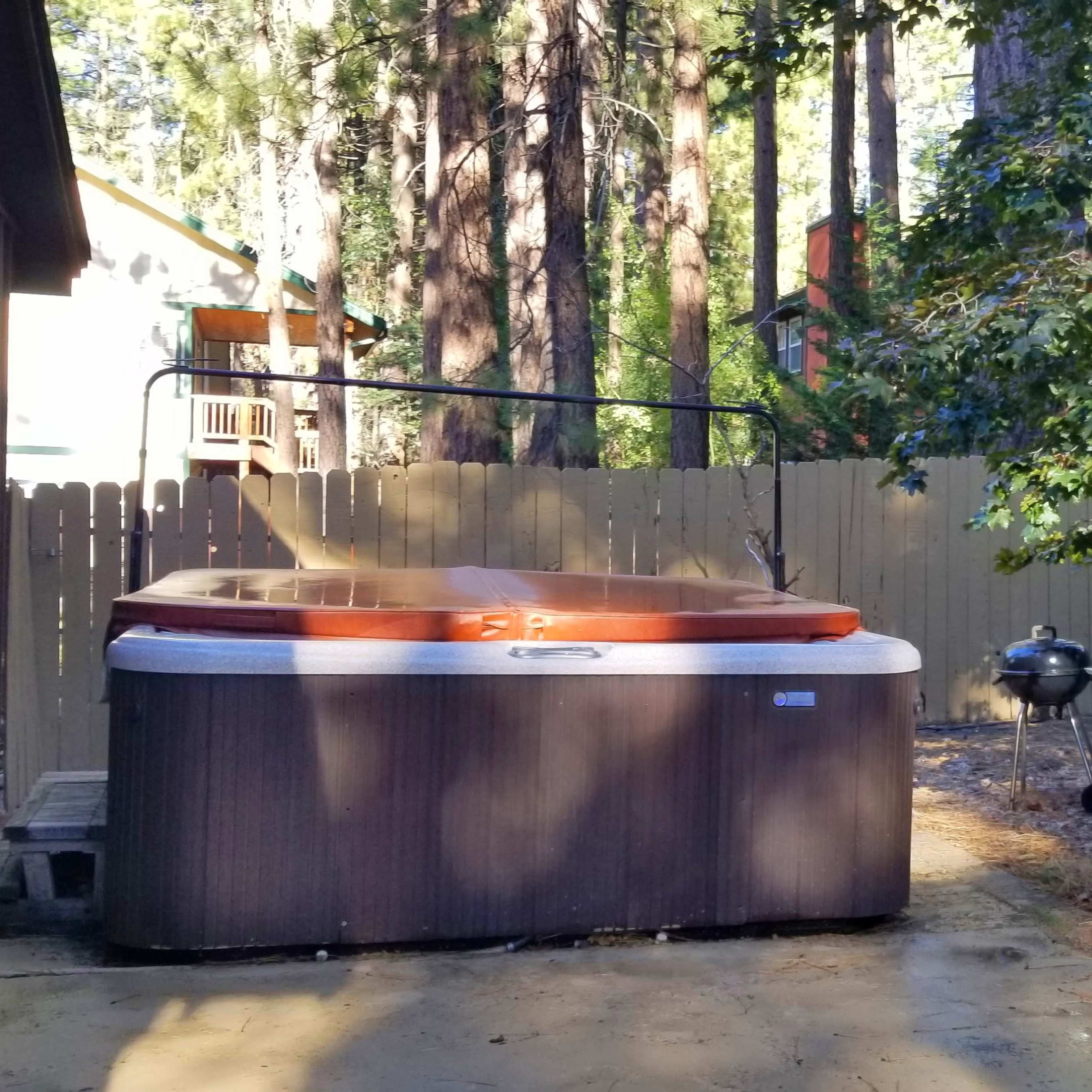 A hot tub next to a barbecue grill and a table with a chair in a wooded backyard.