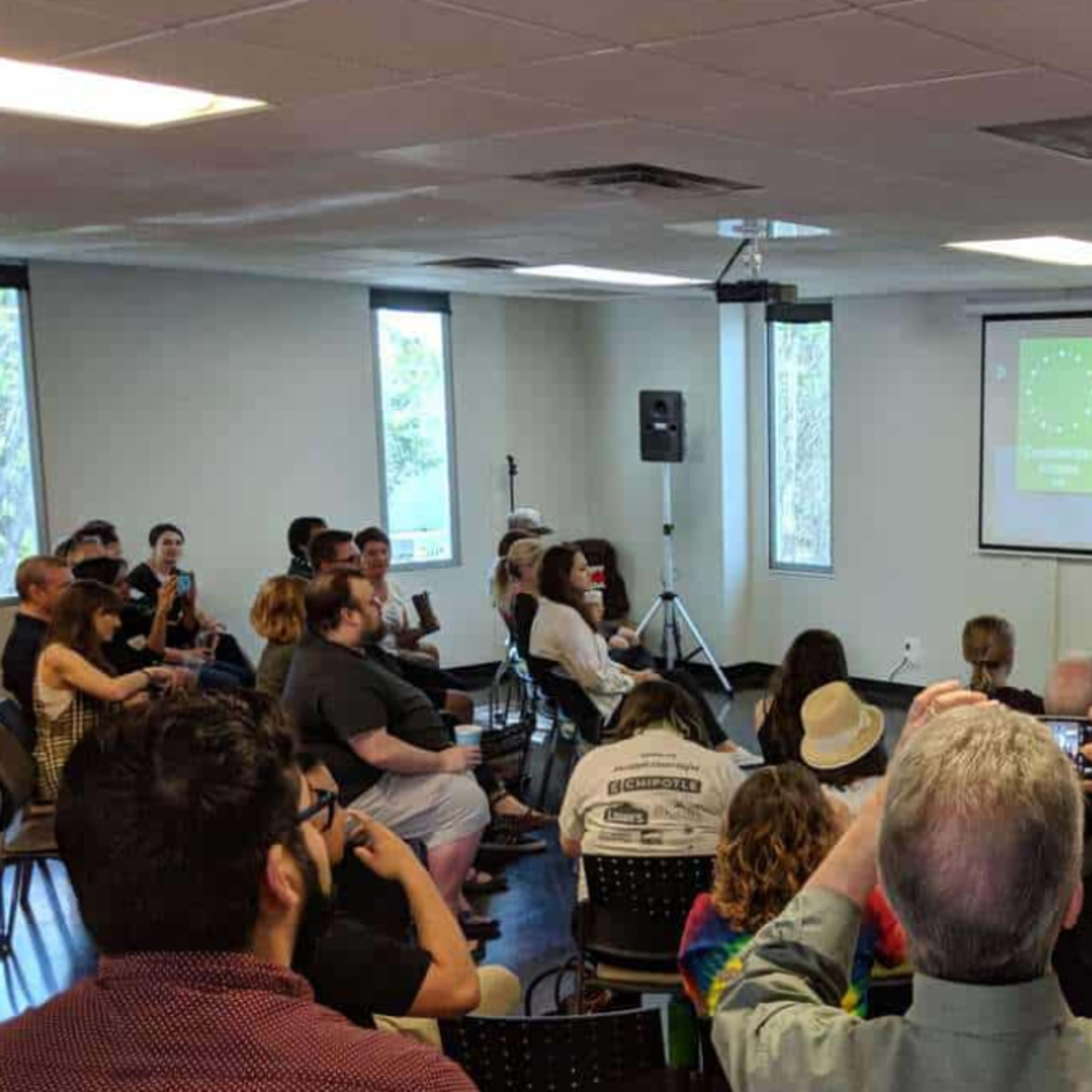A group of people is seated in a conference room while two presenters speak at the front, with a large screen displaying the word "Good Morning" and a hashtag.