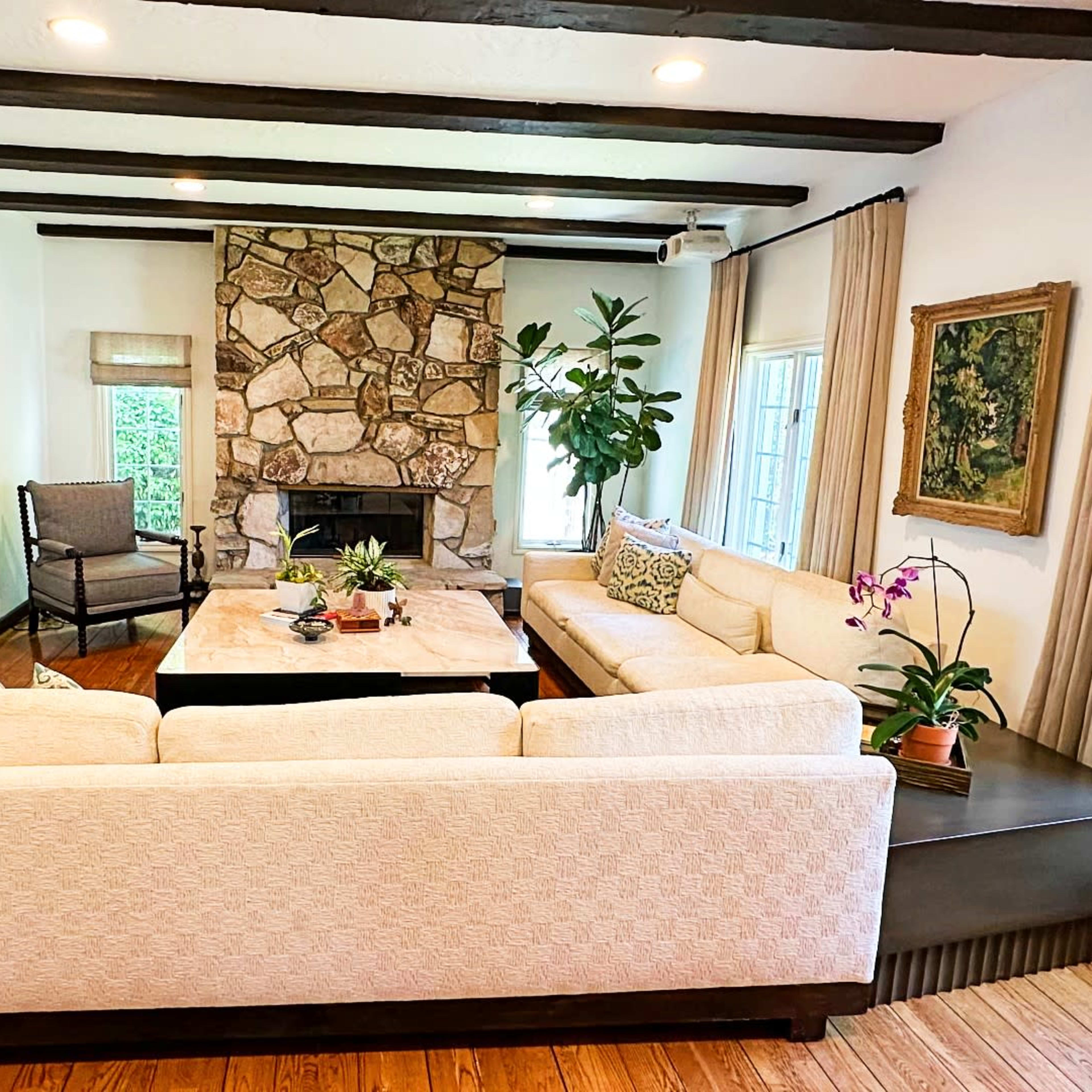 The living room features a large, light-colored sectional sofa facing a stone fireplace, with a coffee table at the center and windows providing natural light.