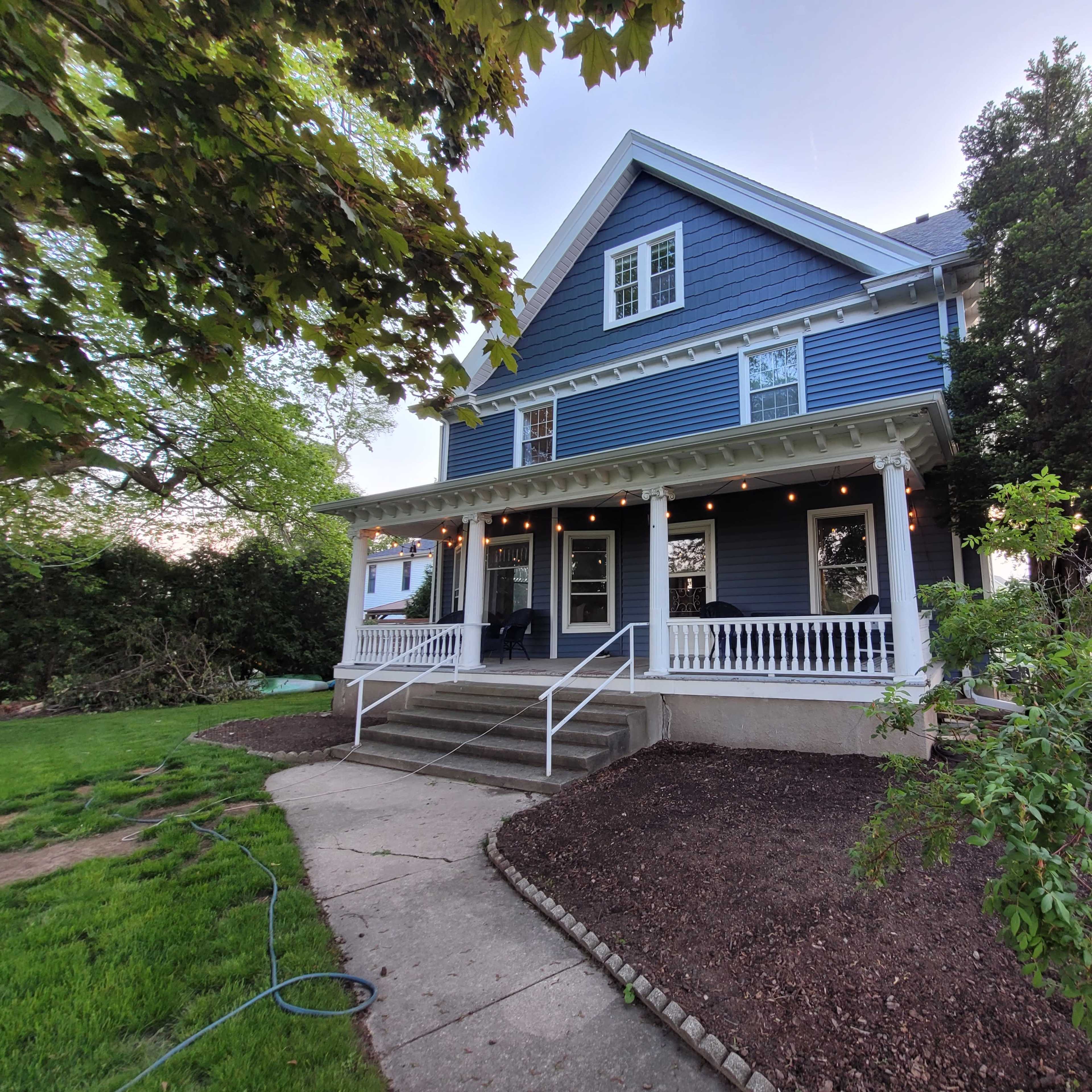 A three-story blue house with a front porch is surrounded by greenery and features outdoor lighting.