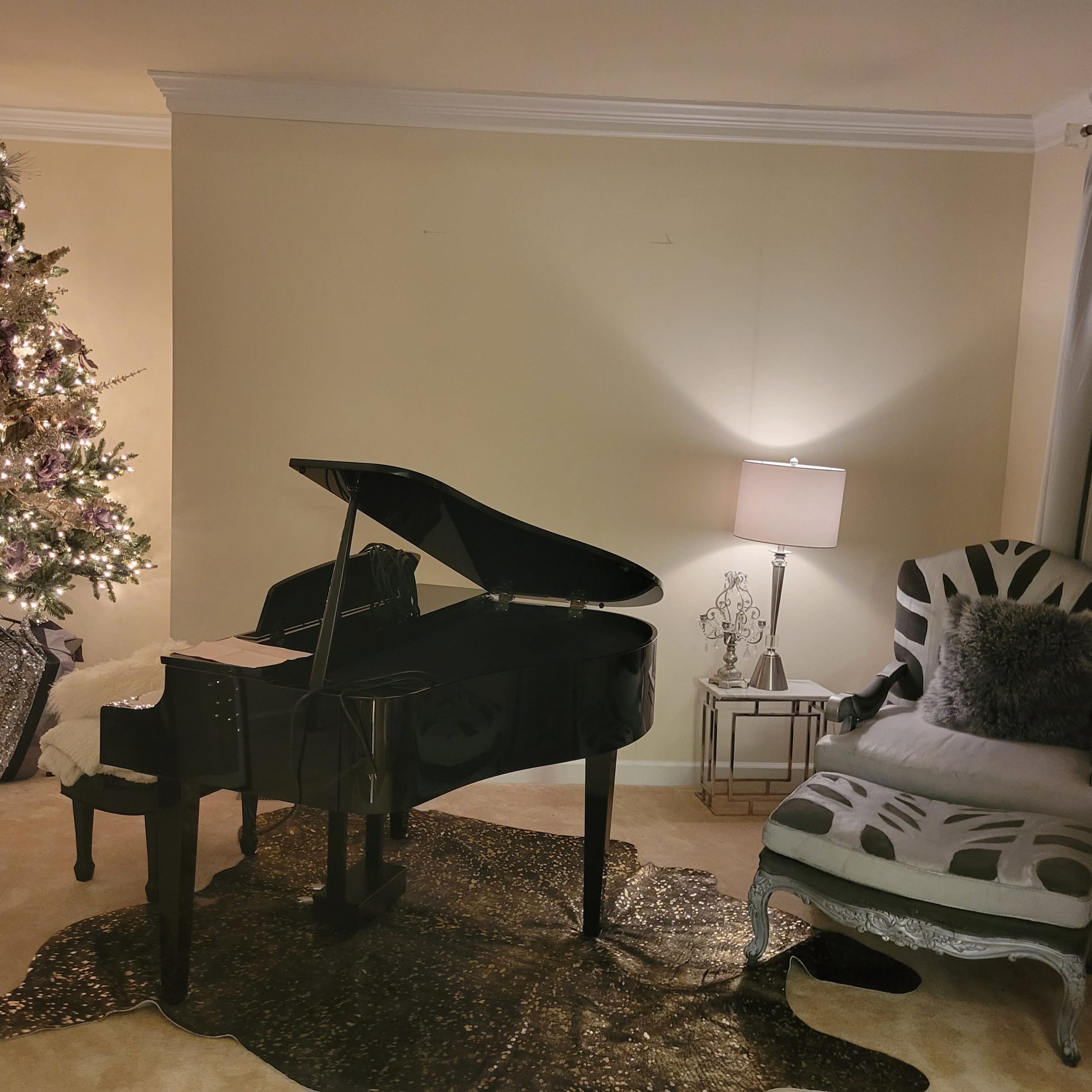 A grand piano sits next to a Christmas tree decorated with lights, while a lamp and an upholstered chair are positioned nearby in a softly lit room.