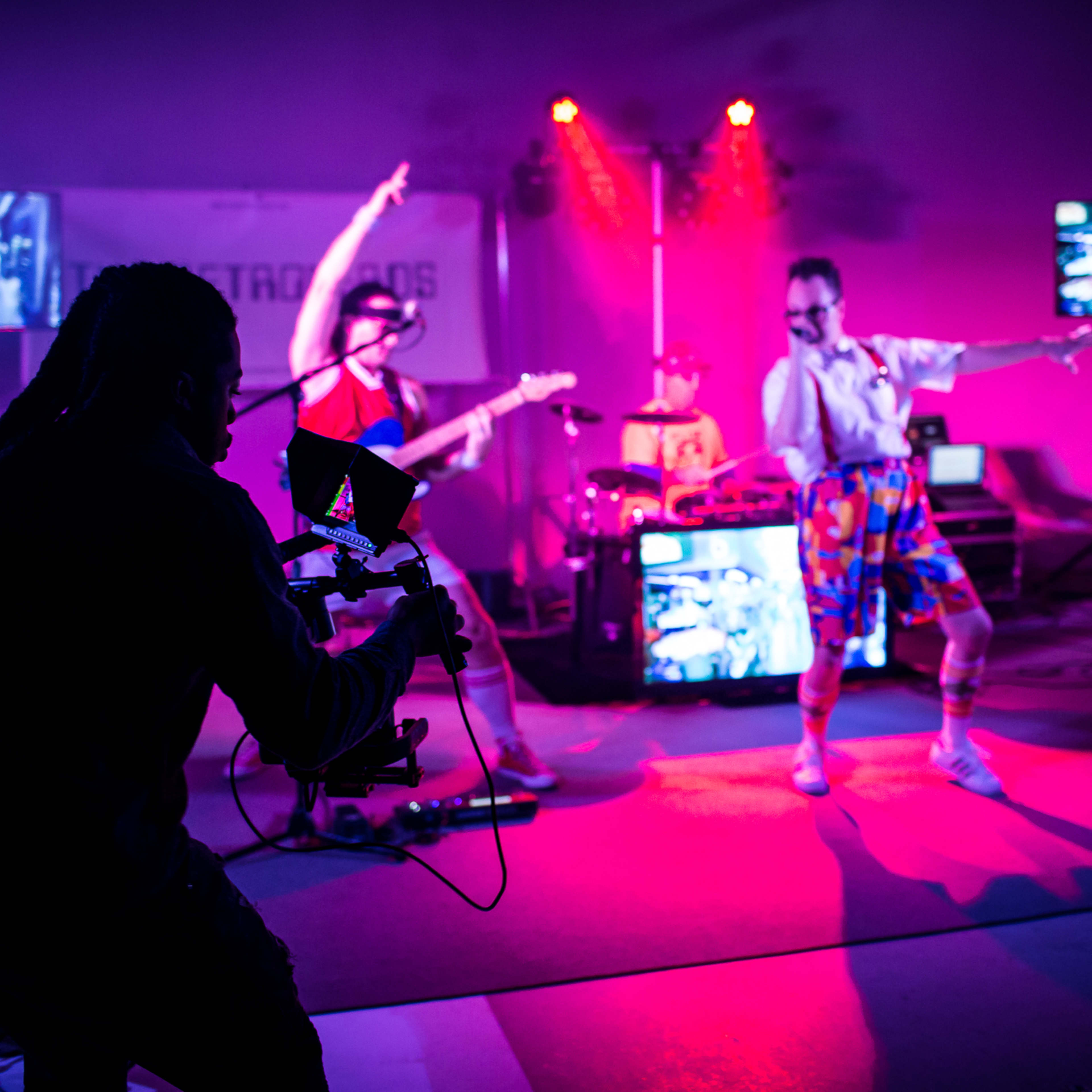 A camera operator captures a band performing on stage, surrounded by colorful lights and video screens displaying their performance.