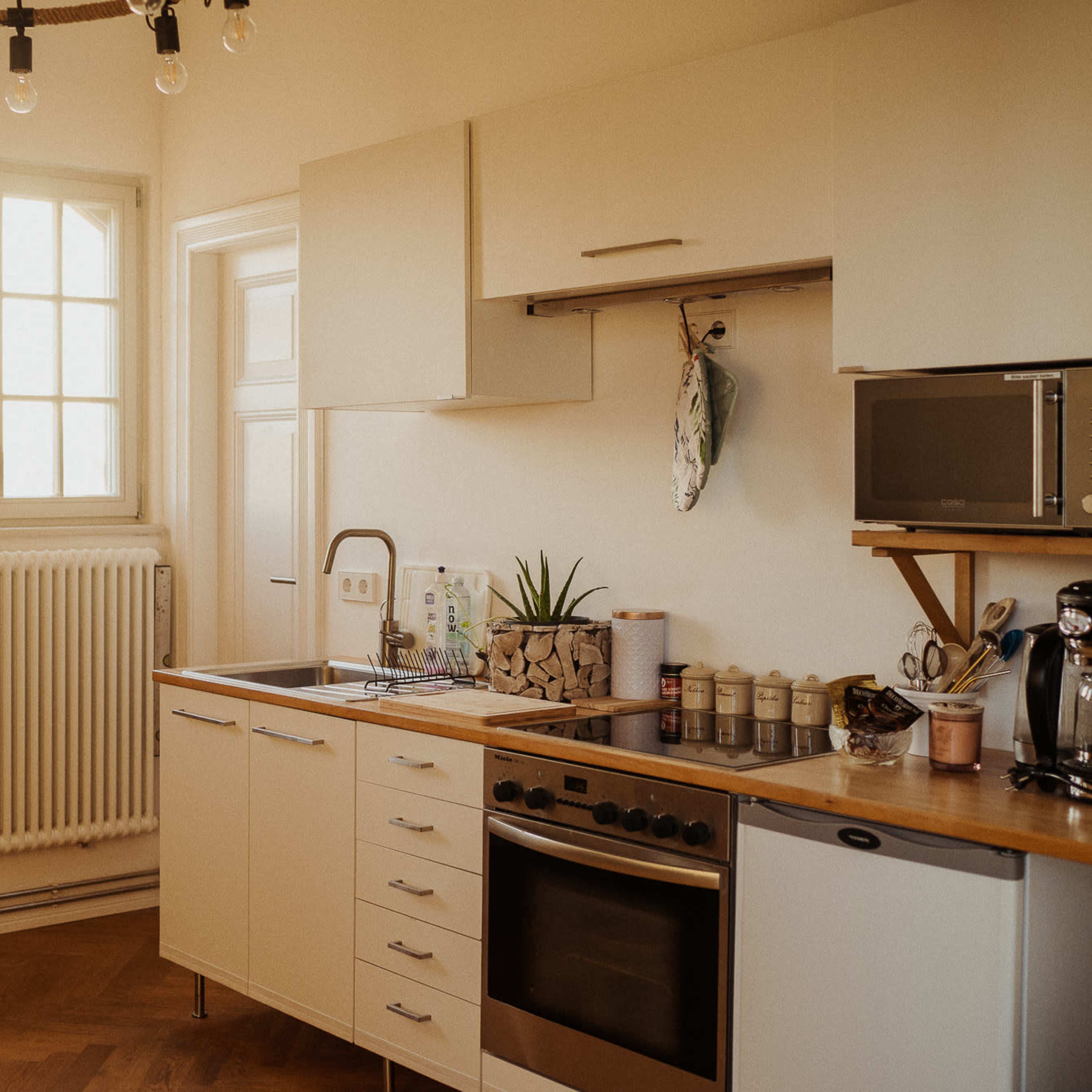 A modern kitchen features a sink, oven, and microwave, with natural light streaming through a window and a radiator along the wall.