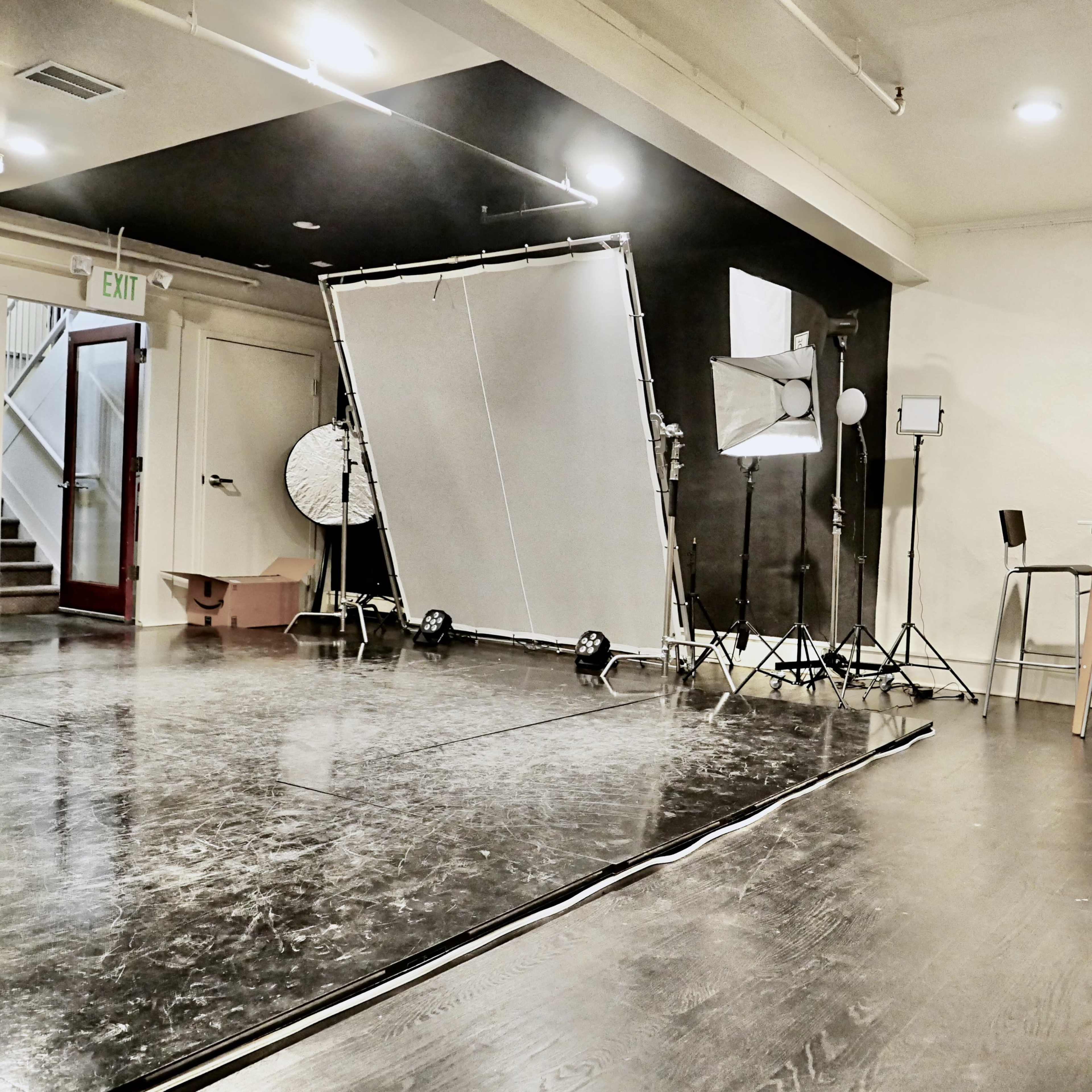 An empty studio space with a large backdrop, lighting equipment, and a table with chairs set against a black wall.