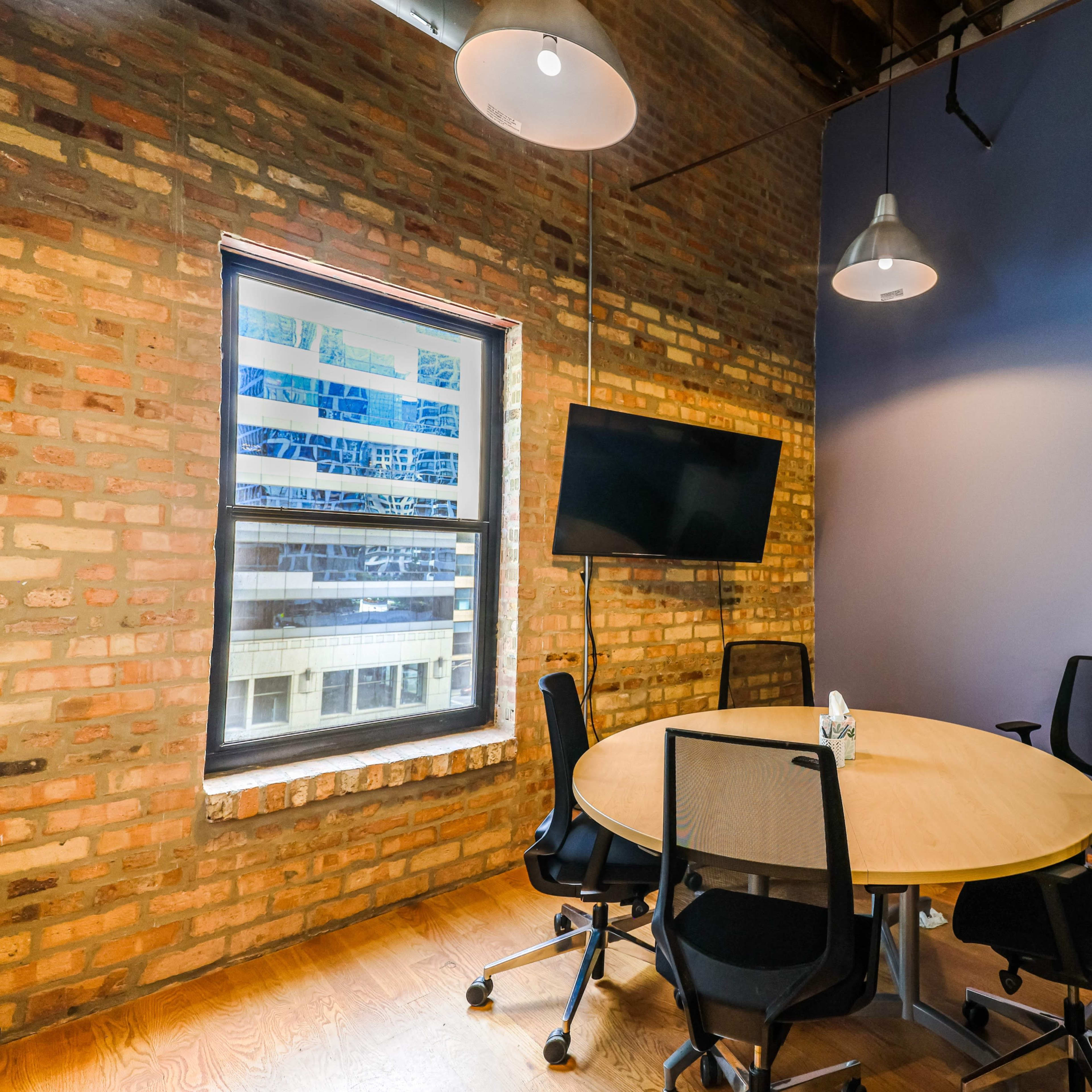 A small conference room with a round table, four chairs, a wall-mounted television, and a filing cabinet, featuring exposed brick and purple walls.