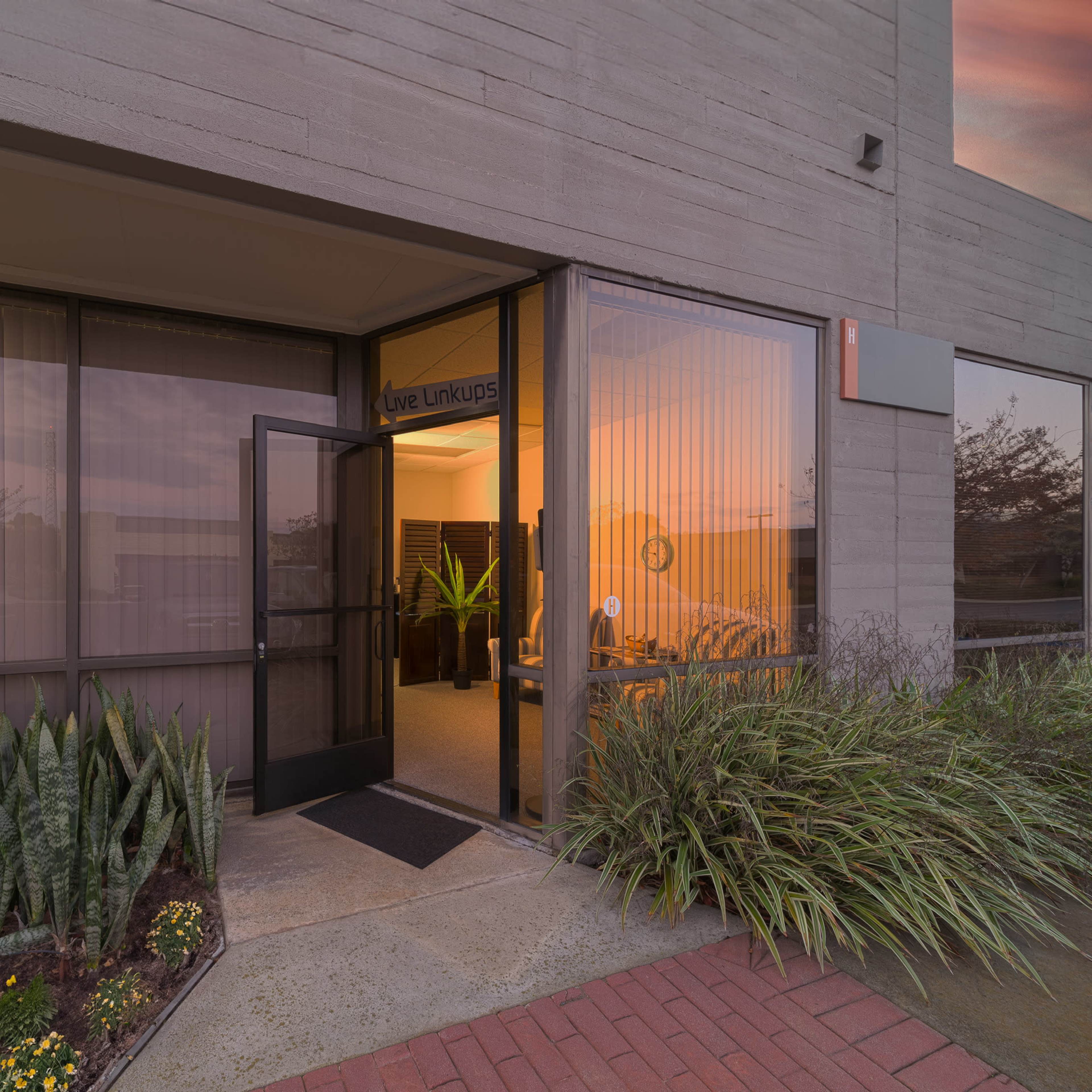 A modern office entrance with glass doors, leading into a softly lit interior surrounded by green plants and a brick pathway.