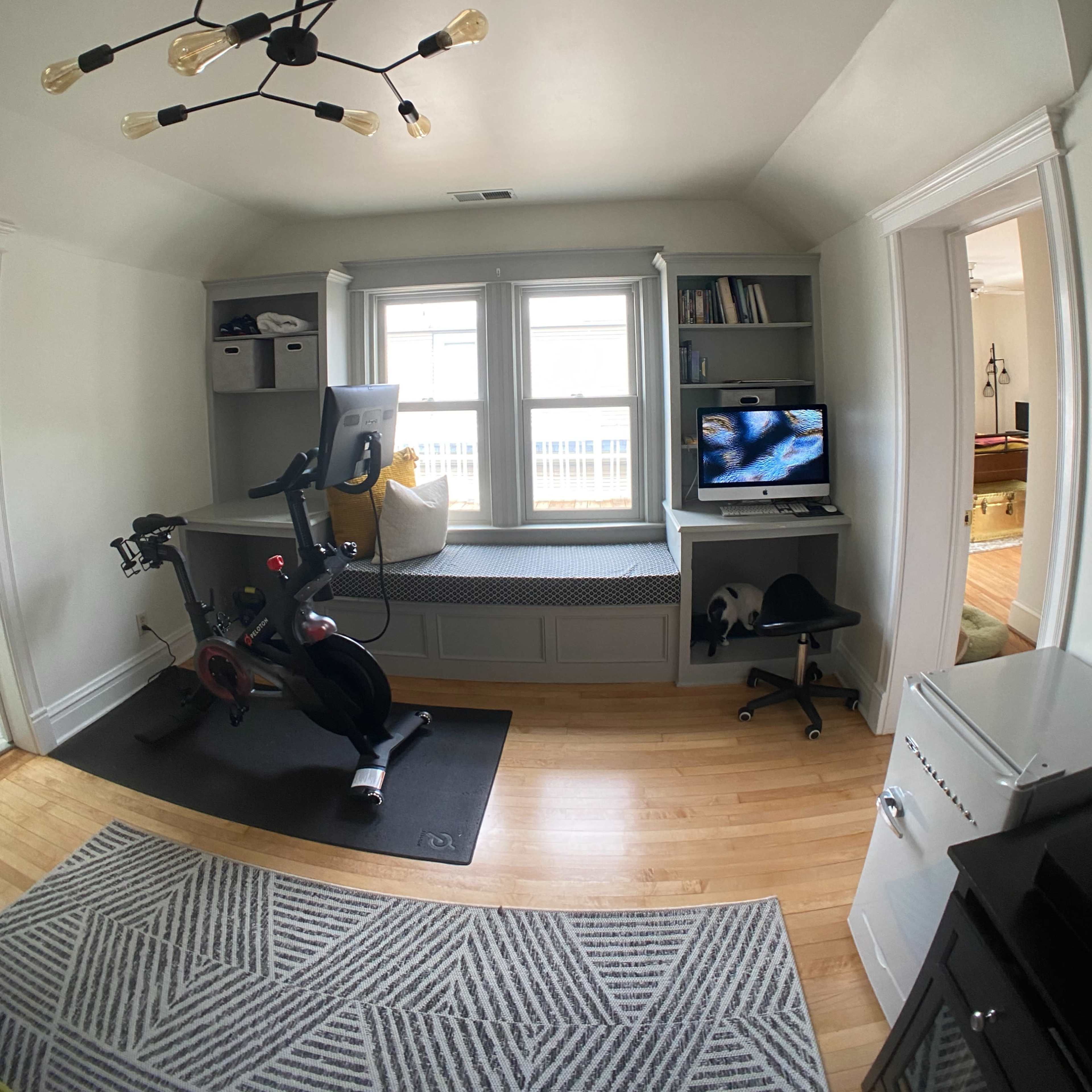 A home office with a stationary exercise bike, a small desk with a monitor, and built-in window seats.