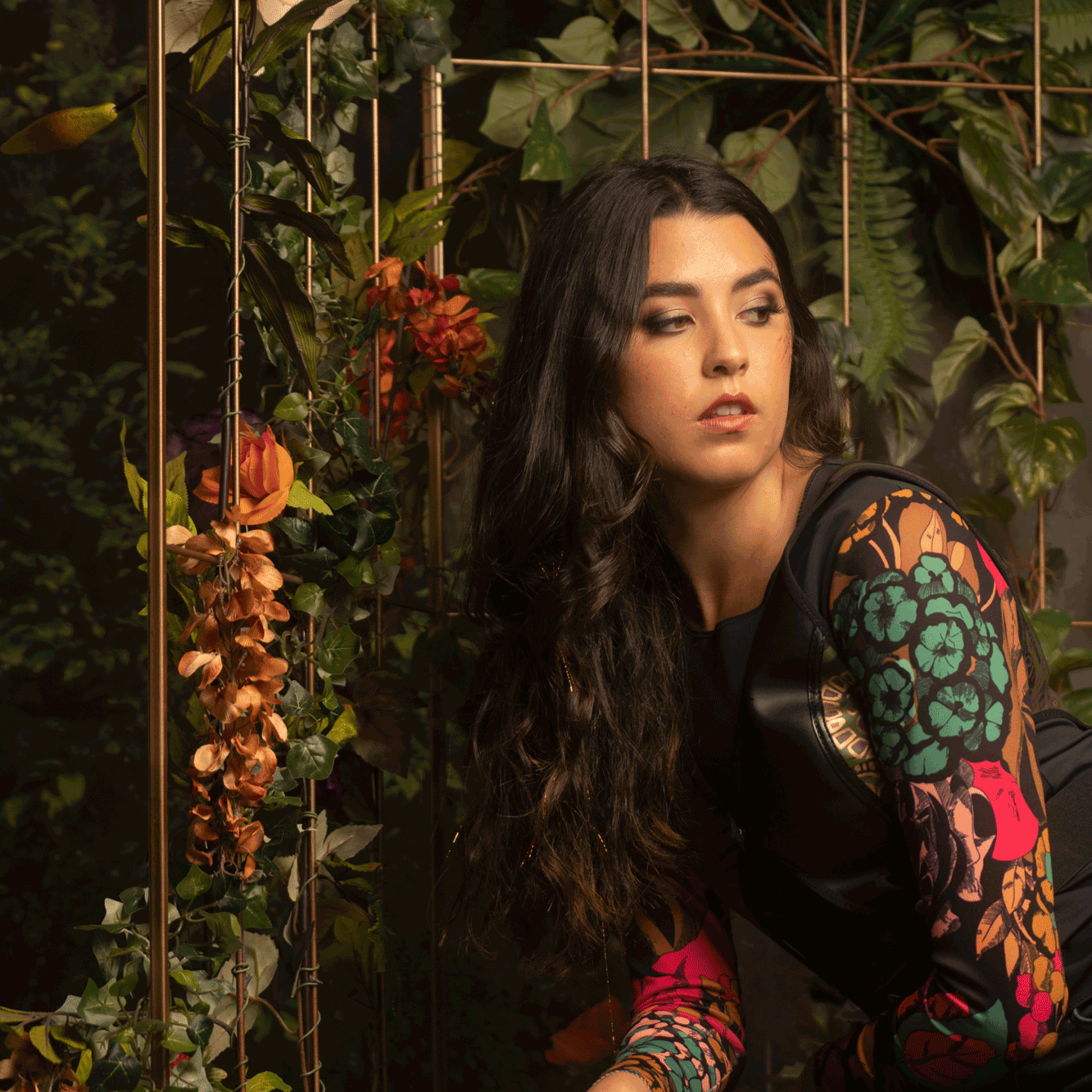 A woman with long, dark hair poses in front of a backdrop of vibrant flowers and greenery.