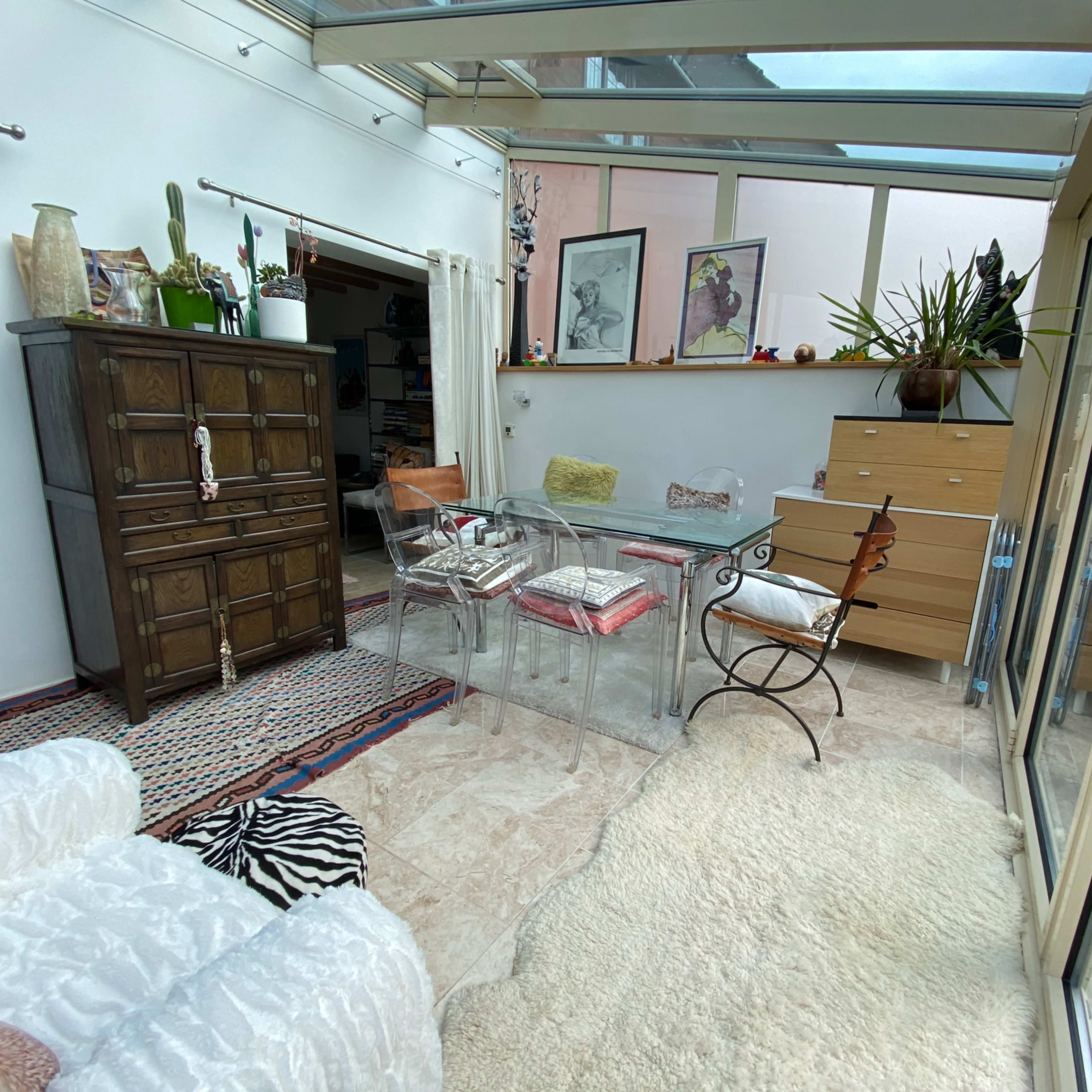 A sunlit room with a glass table surrounded by transparent chairs, a wooden cabinet, and various plants in the background.