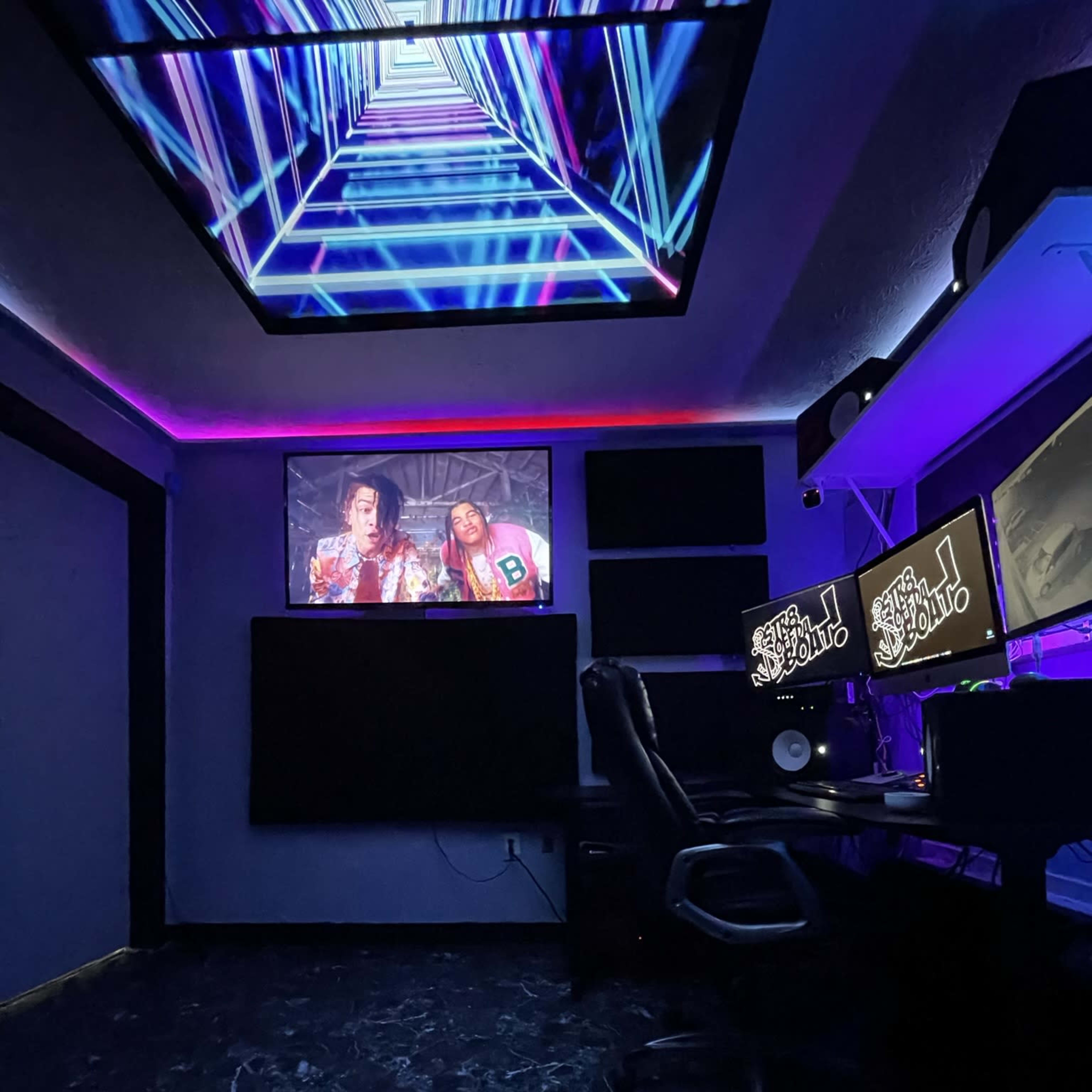 A music studio with a large screen displaying a video, neon lighting, and two computer monitors on a desk.