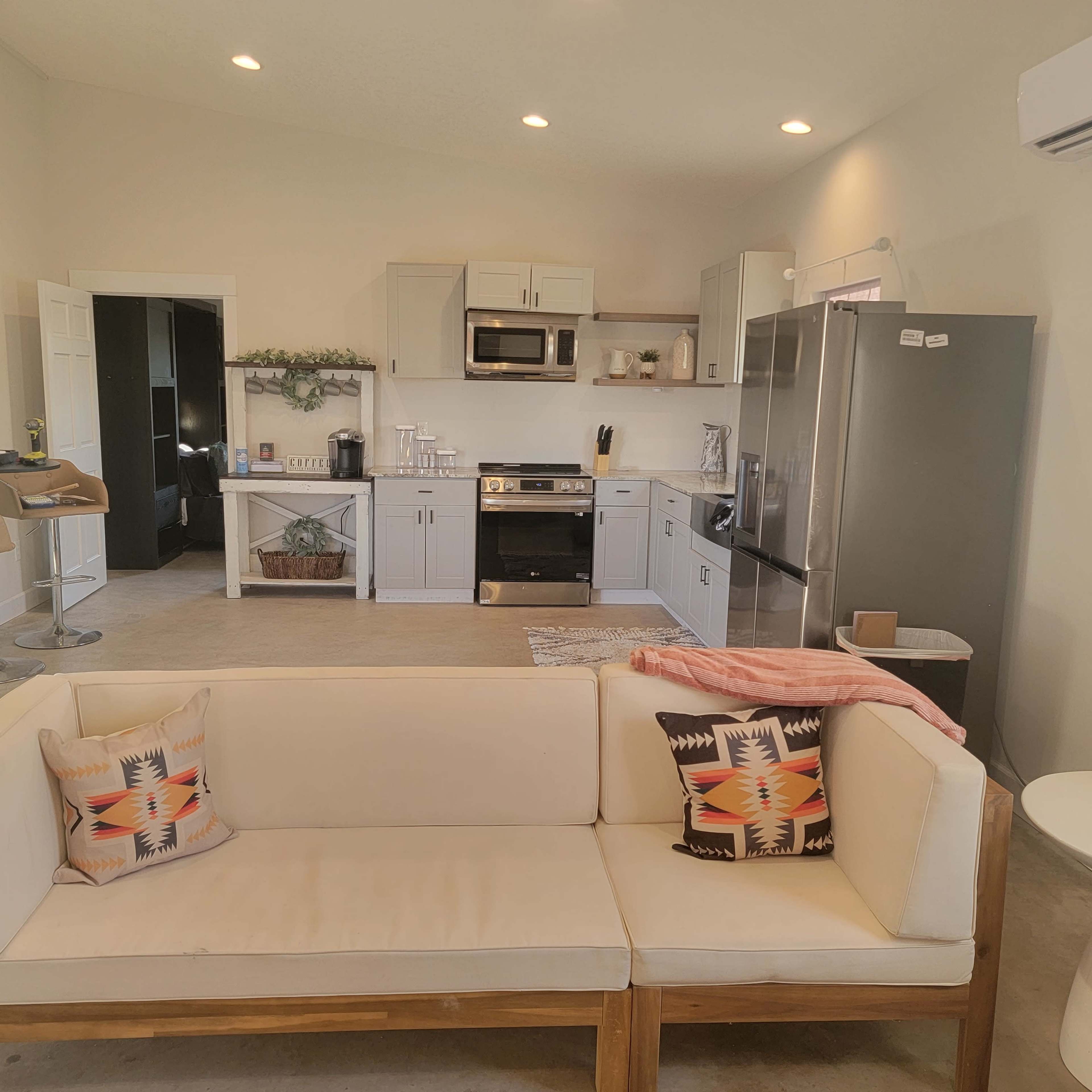 A modern kitchen and living area featuring a beige sofa with patterned cushions, a dining counter with stools, and stainless steel appliances.