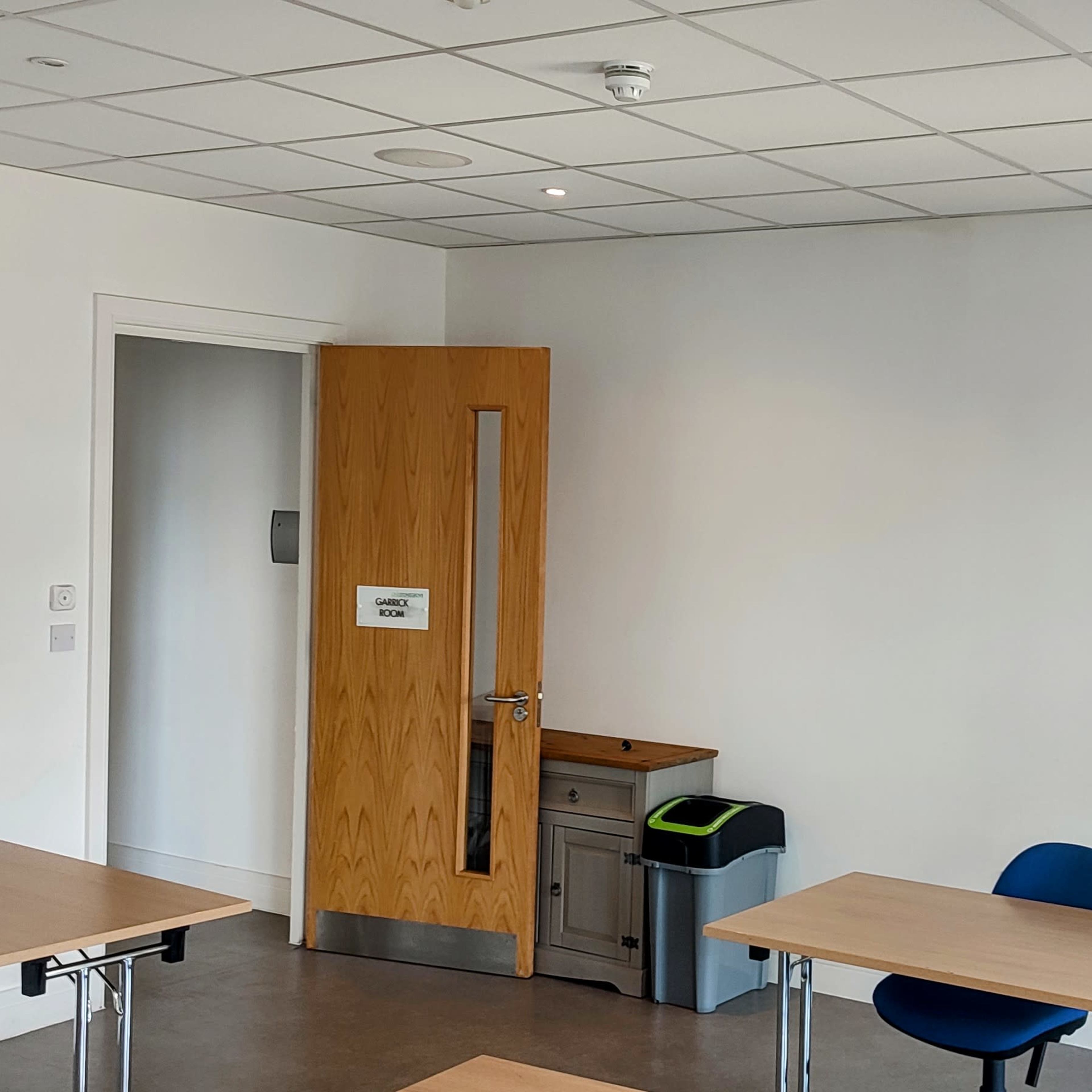 A bright, empty meeting room features two wooden tables, a blue chair, a television mounted on the wall, a door with a window, and a plant in a pot beside a waste bin.