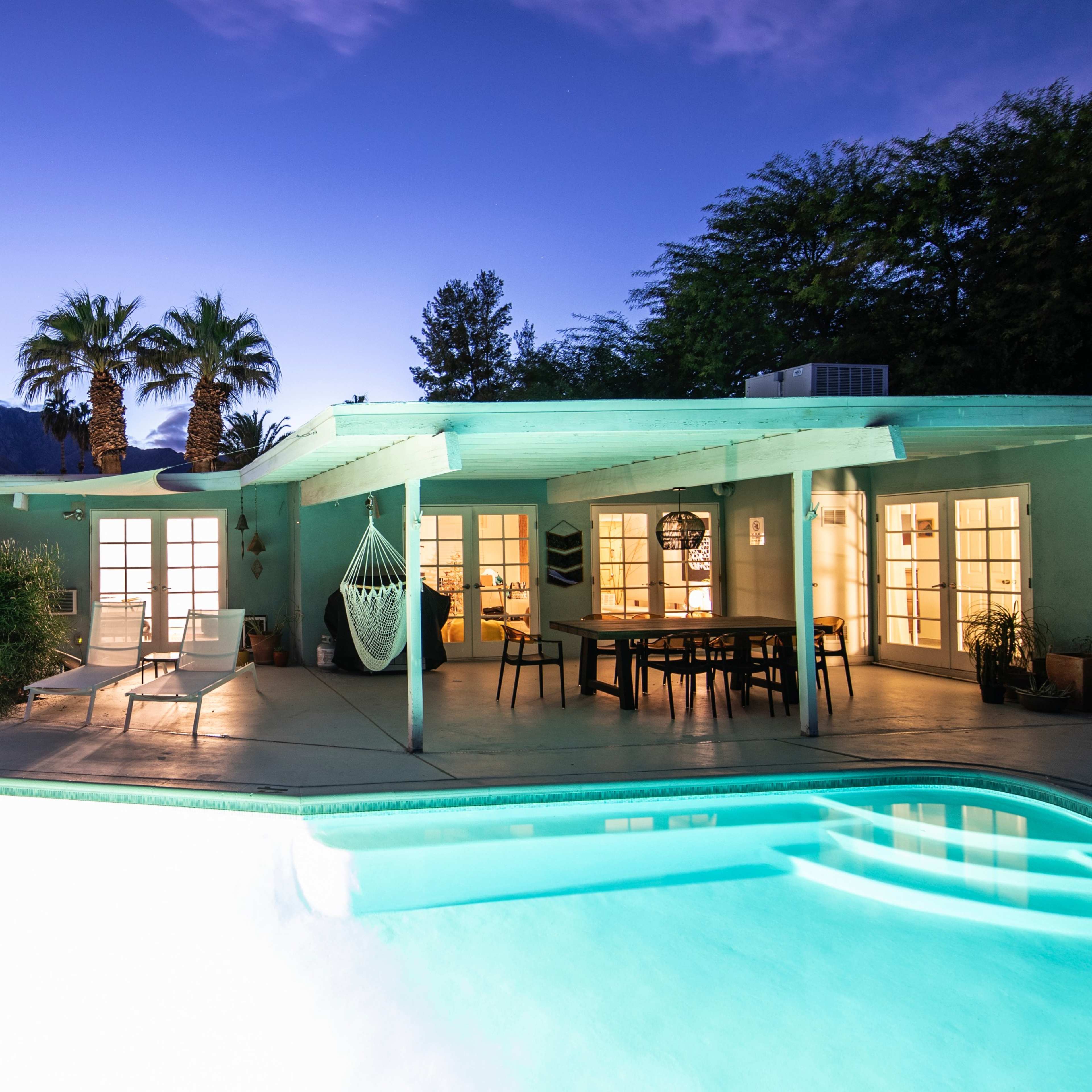 A poolside view of a modern home at dusk, with a clear sky and mountains in the background.