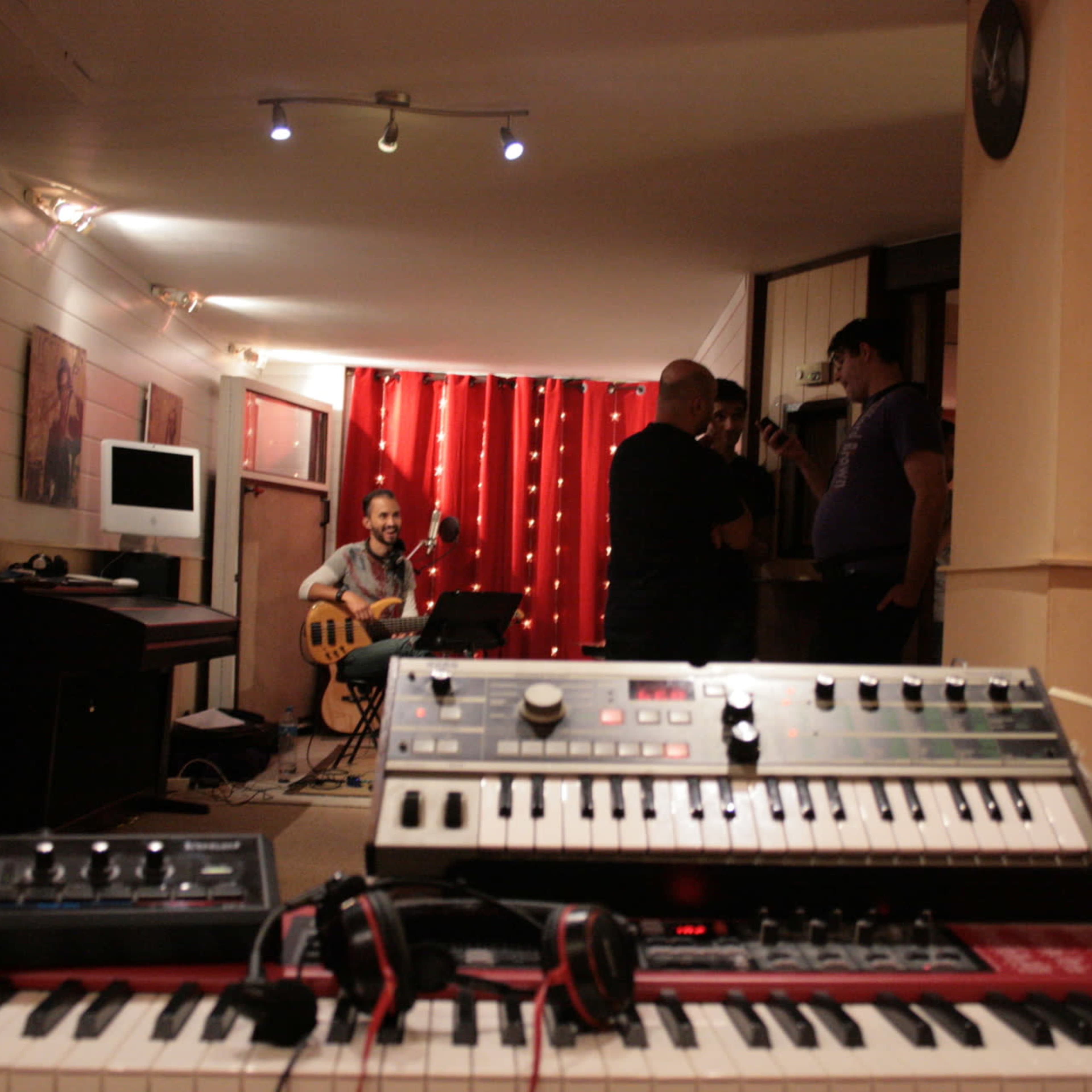 A musician plays a guitar while a group of people converse in a dimly lit room featuring various musical instruments.