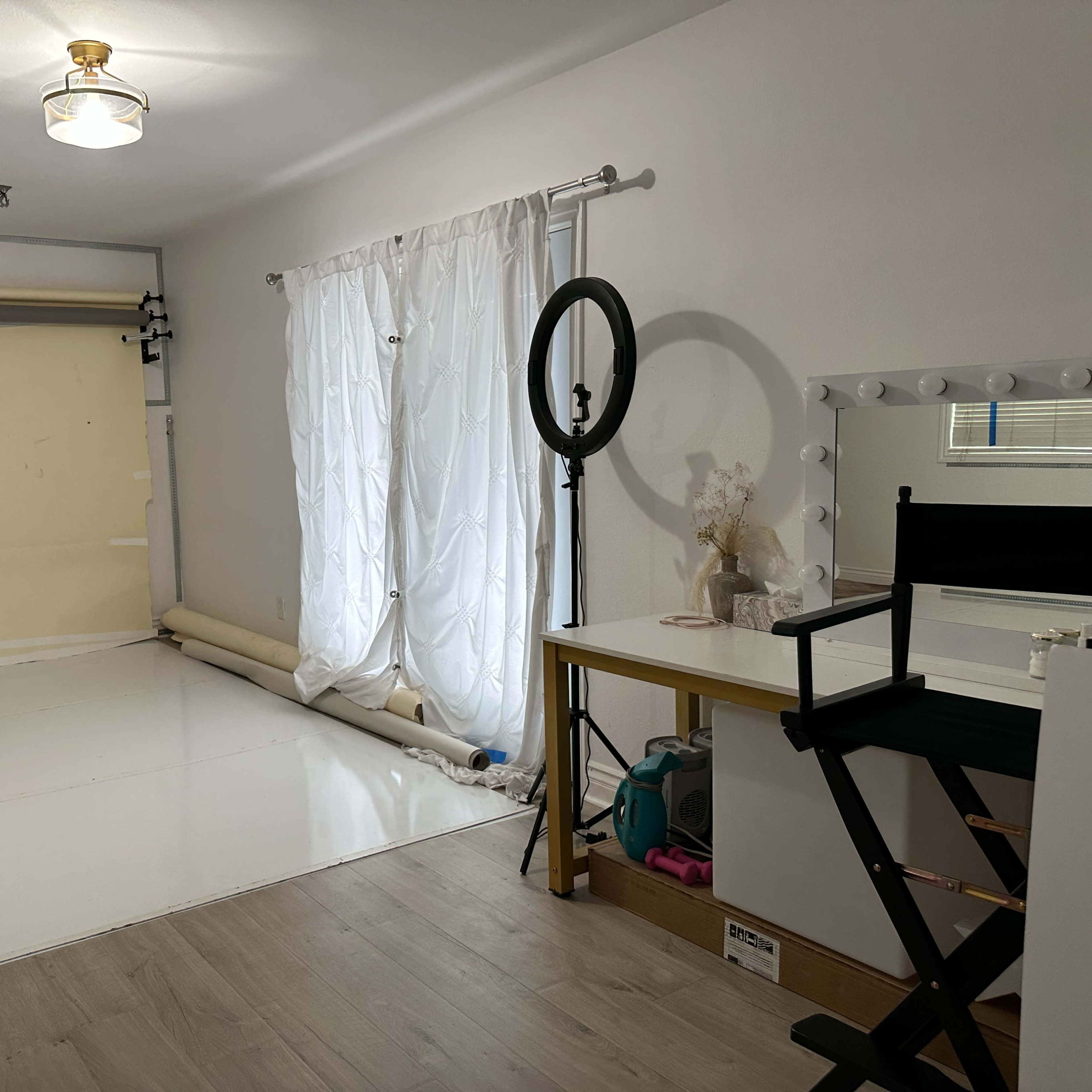 A studio space featuring a light-colored floor, a large window with white curtains, a backdrop setup, a makeup mirror with lighting, and a black director's chair.