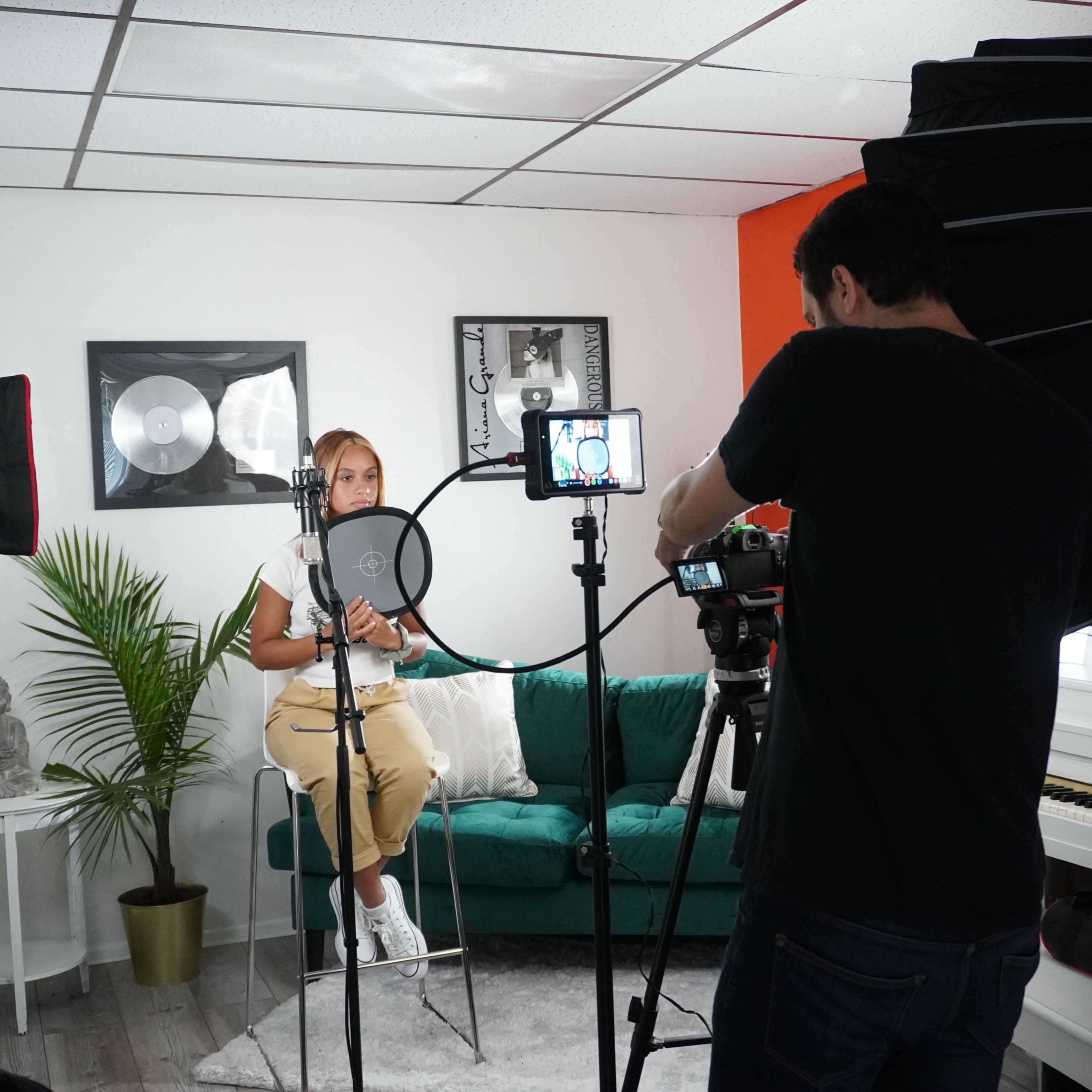 A woman sits on a teal couch in a studio setting while a man operates a camera with bright lights positioned around them.