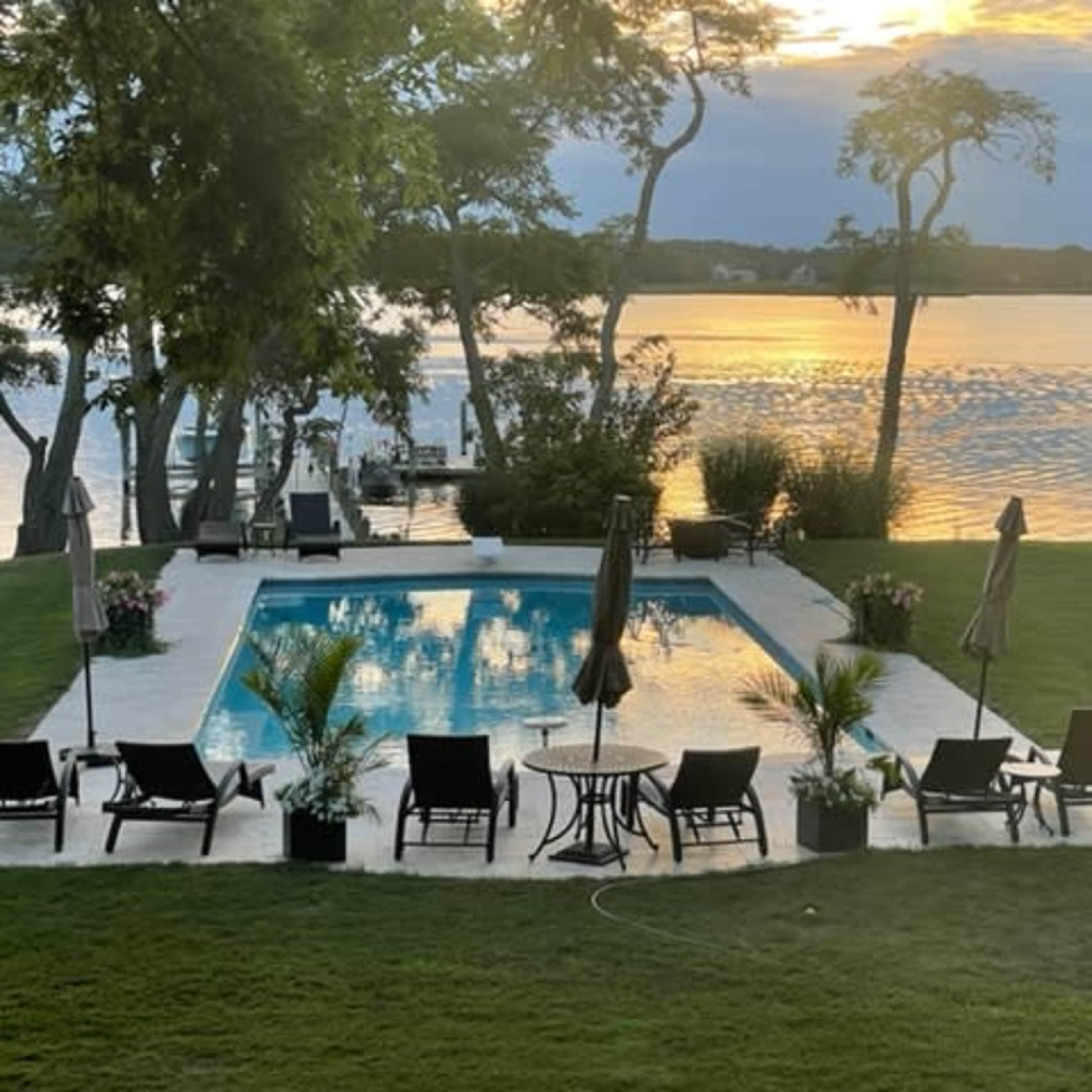 A swimming pool surrounded by lounge chairs and umbrellas, set against a lake with a sunset reflecting on the water.