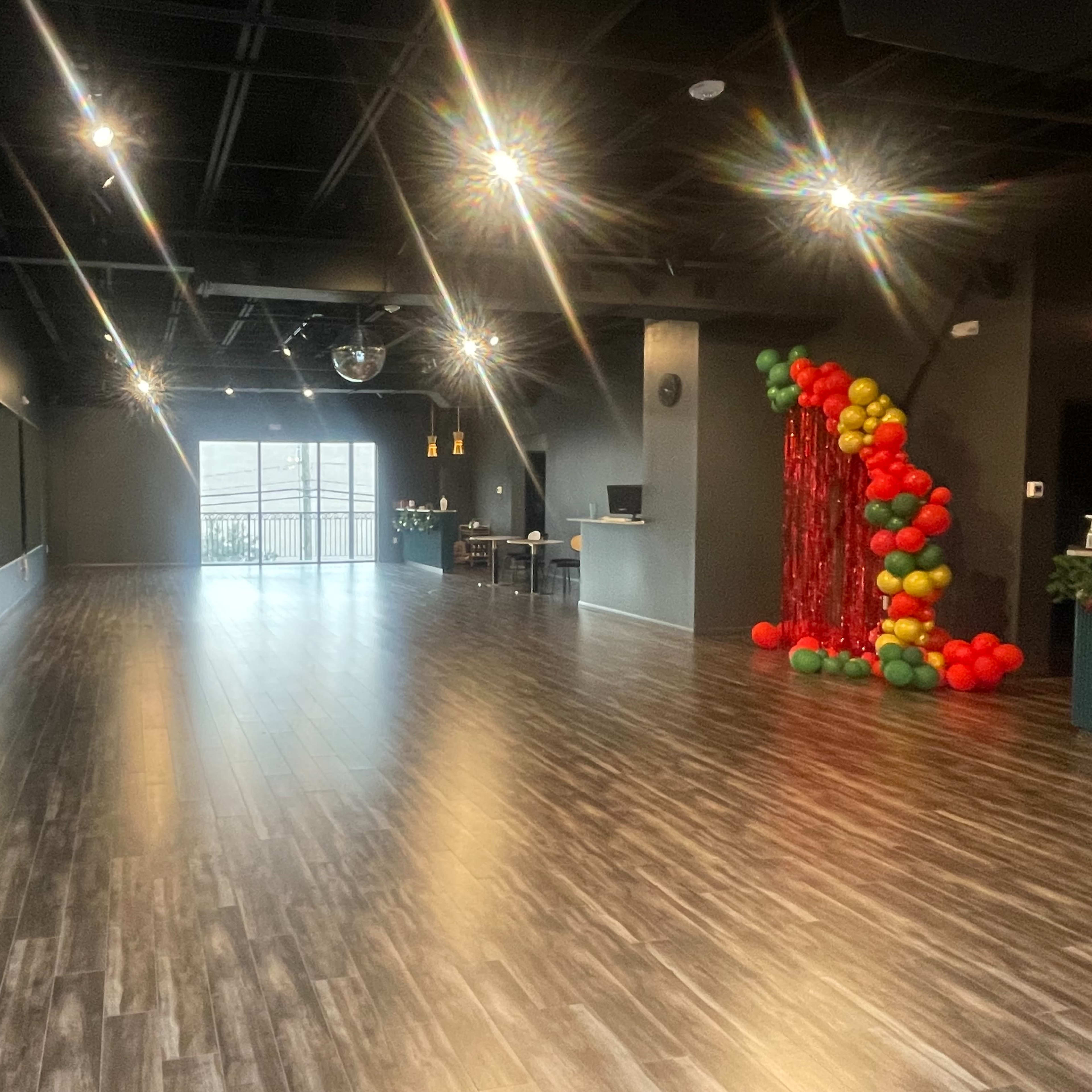 A spacious, well-lit studio features mirrored walls, polished wooden floors, and a decorative arrangement of colorful balloons on one side.