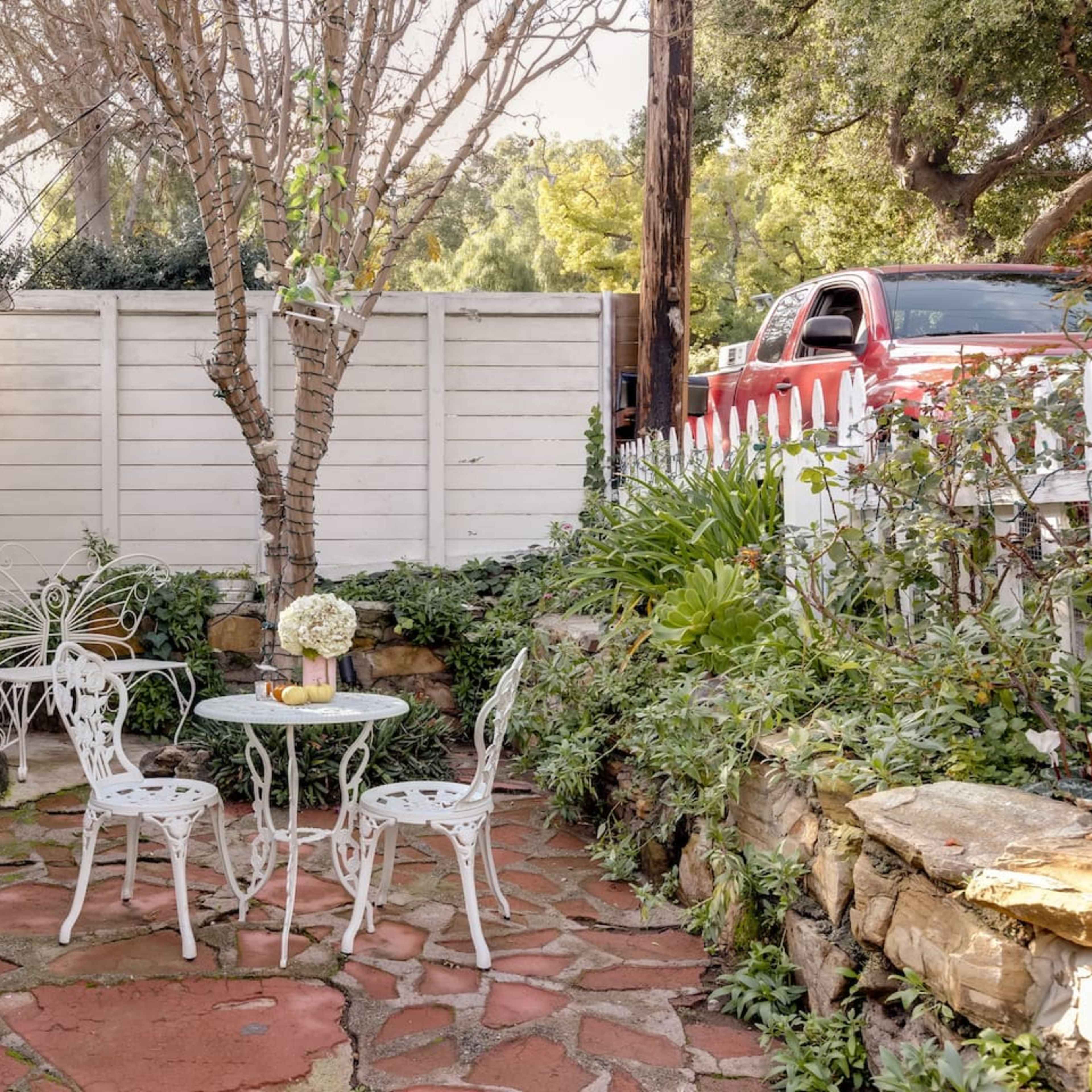 A small stone-paved patio features a round table and four white chairs, surrounded by greenery and a bare tree, with a house and a white fence in the background.