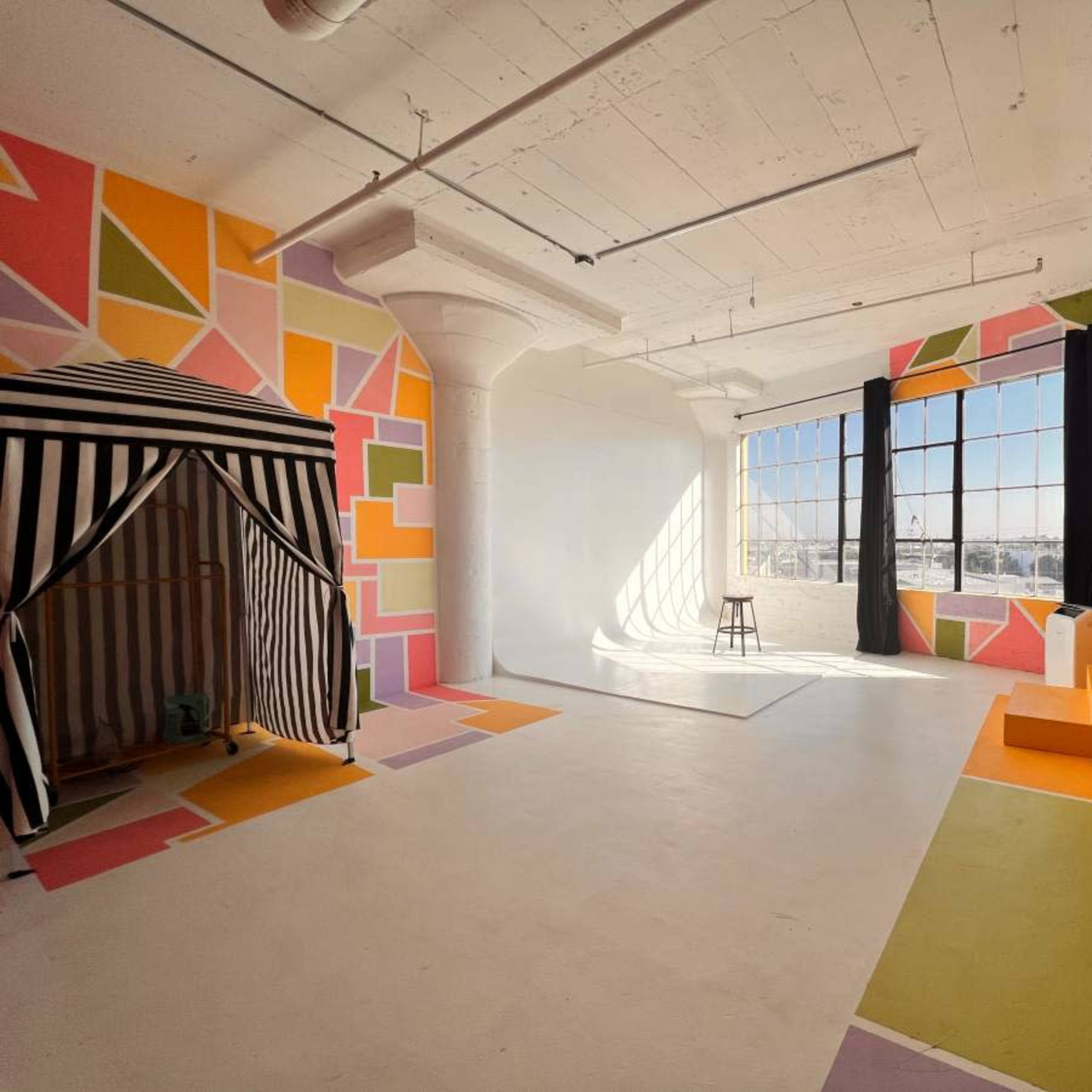 A brightly colored studio space featuring geometric wall patterns, a black and white striped tent, a stool, and various shaped structures in orange and yellow.