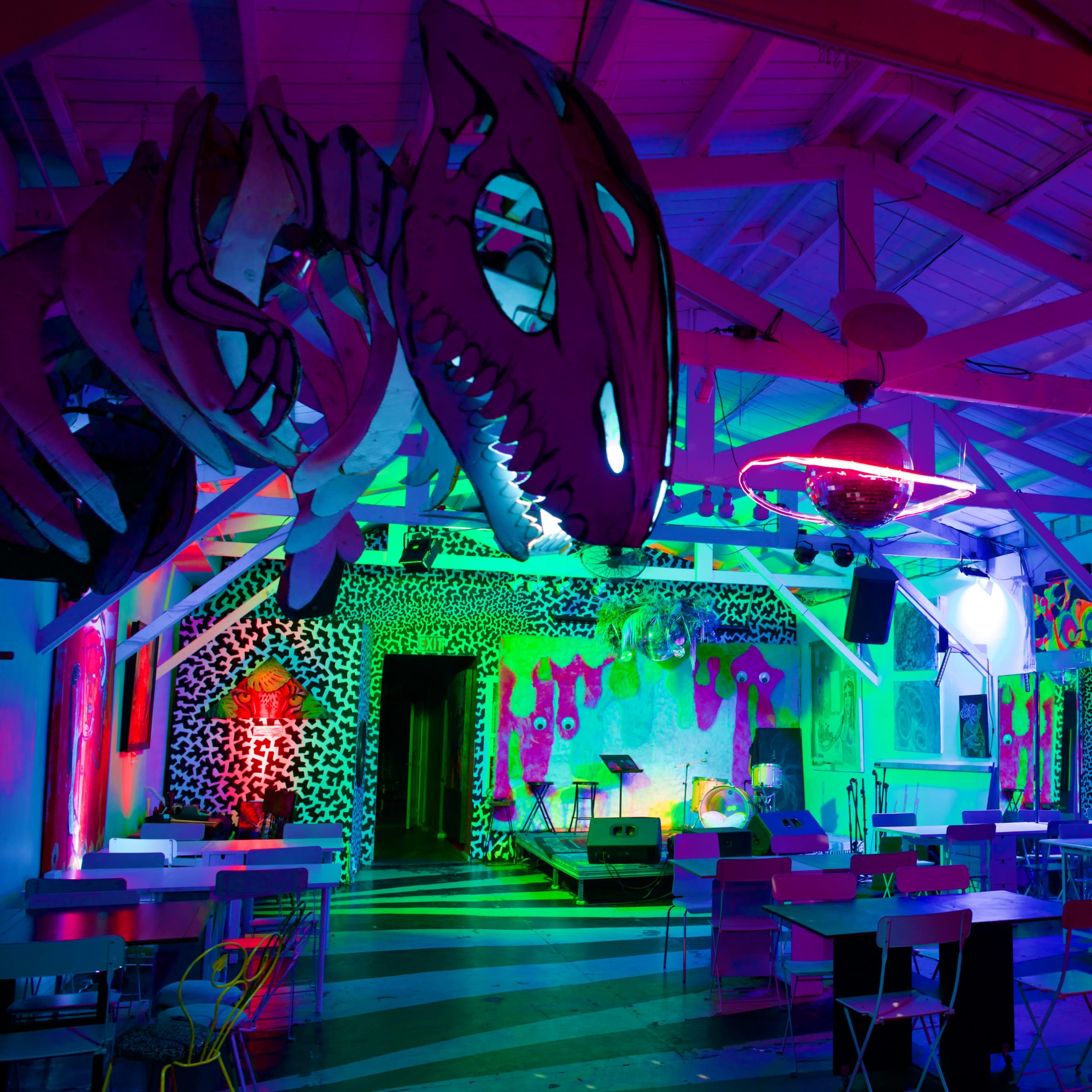 A brightly colored, artistic event space with neon lighting and eclectic decorations, including sculptures and mural-style artwork on the walls.