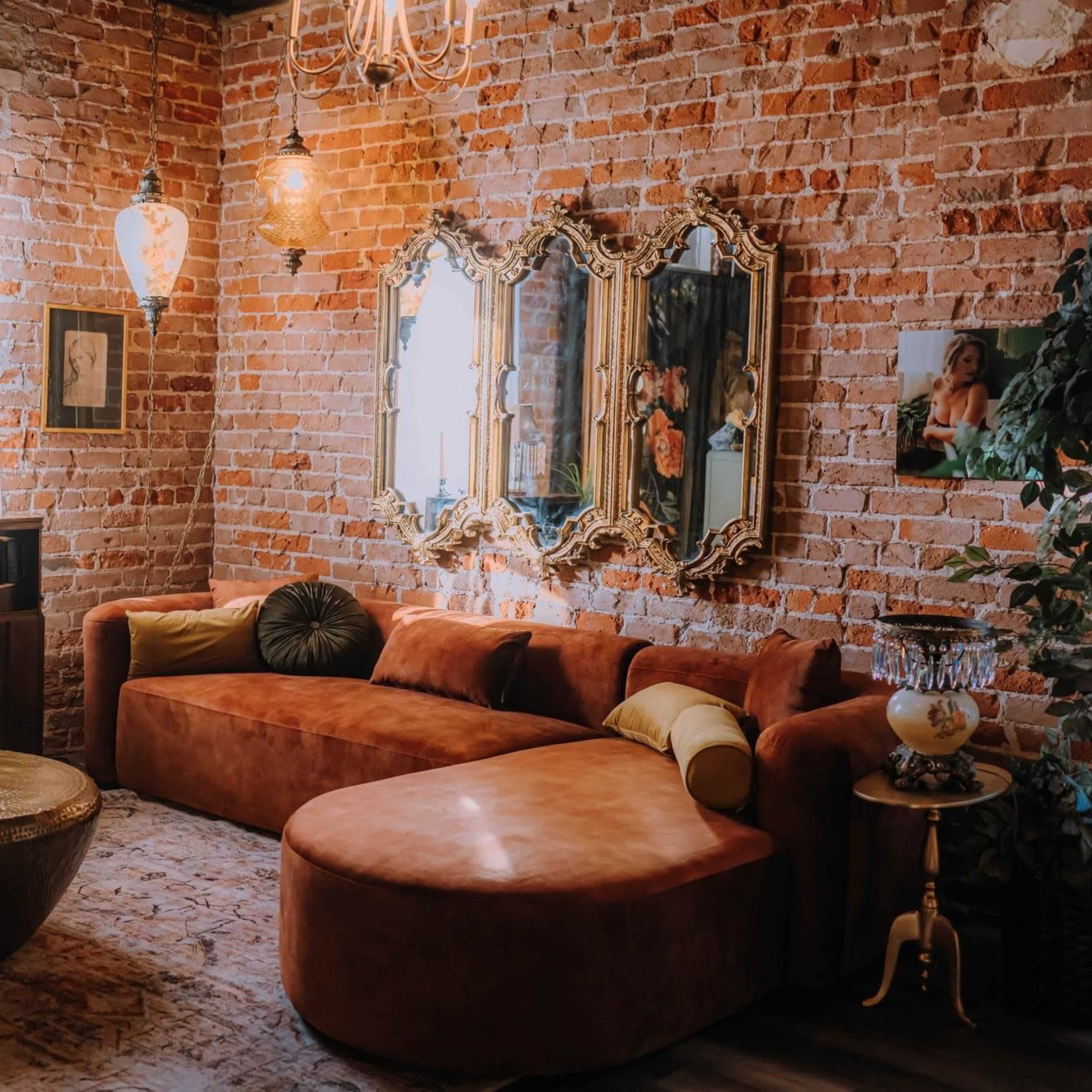 A cozy living room with exposed brick walls, a curved orange sofa, a round coffee table, and elegant mirrors reflecting warm lighting.