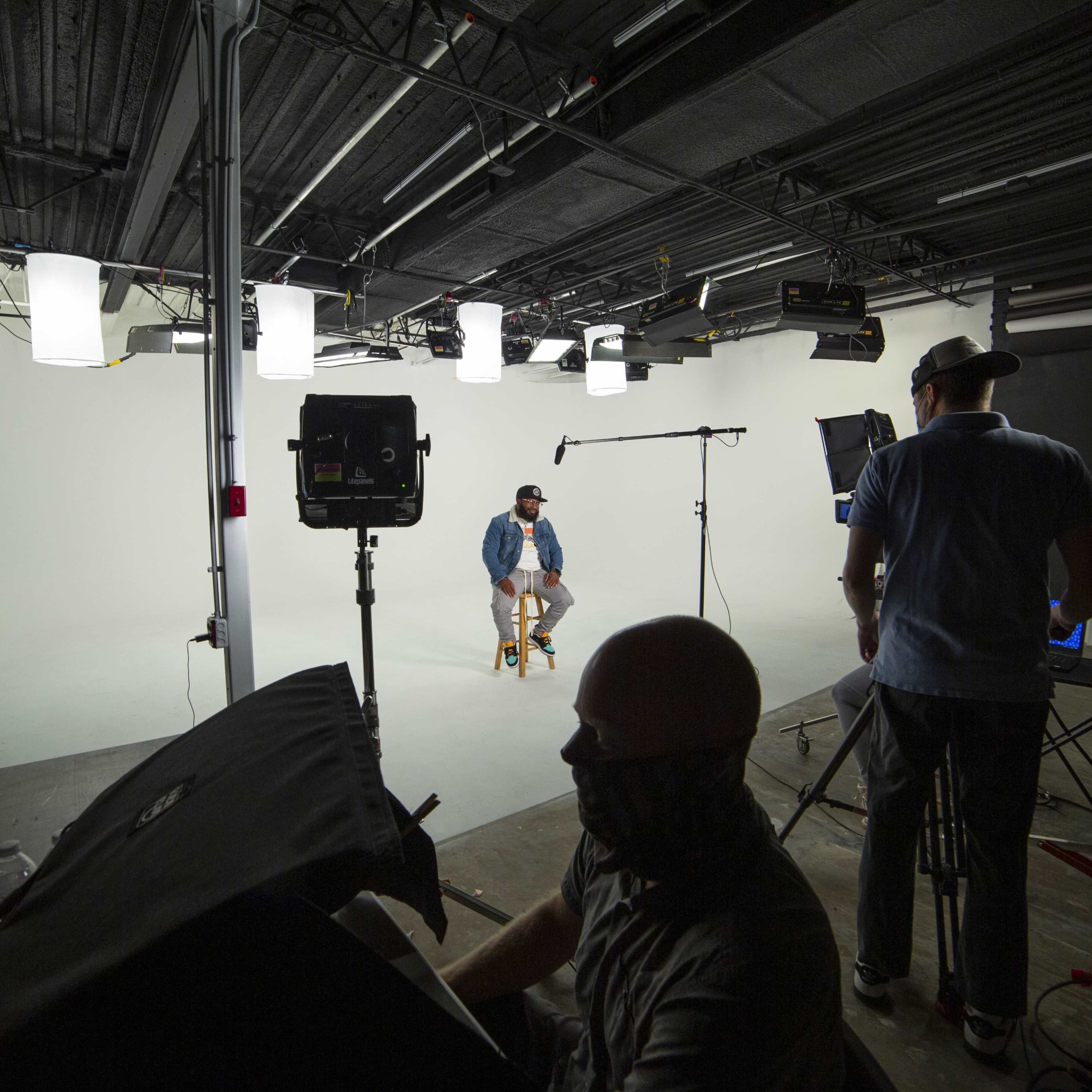 A film studio set is shown with a man sitting on a stool in front of a blank backdrop, while crew members operate cameras and lighting equipment.