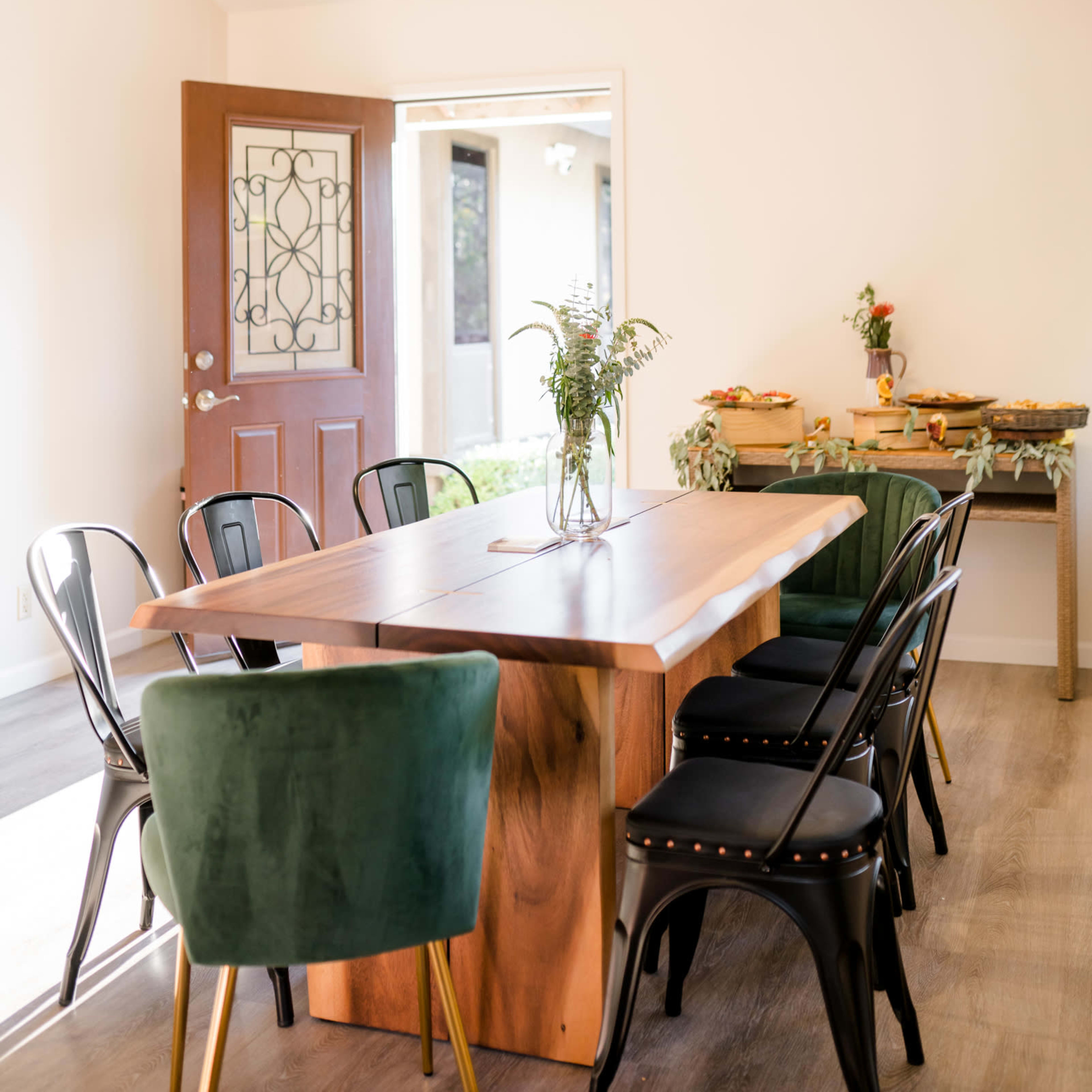 A modern dining area features a wooden table surrounded by black and green chairs, with a decorative plant centerpiece and a doorway leading outside.