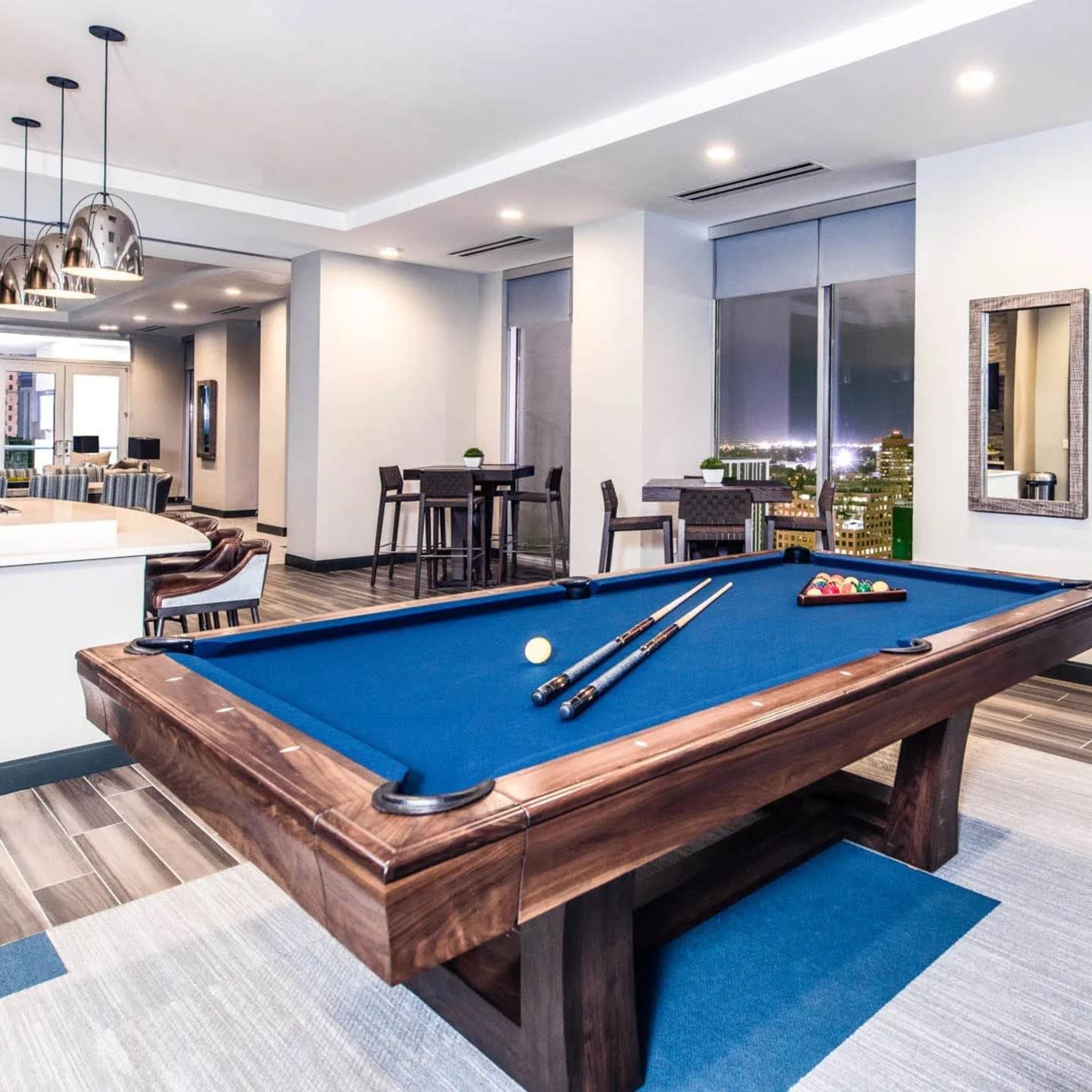 A well-lit game room features a blue pool table with cues and balls prepared for play, surrounded by modern furniture and large windows showing a city skyline.