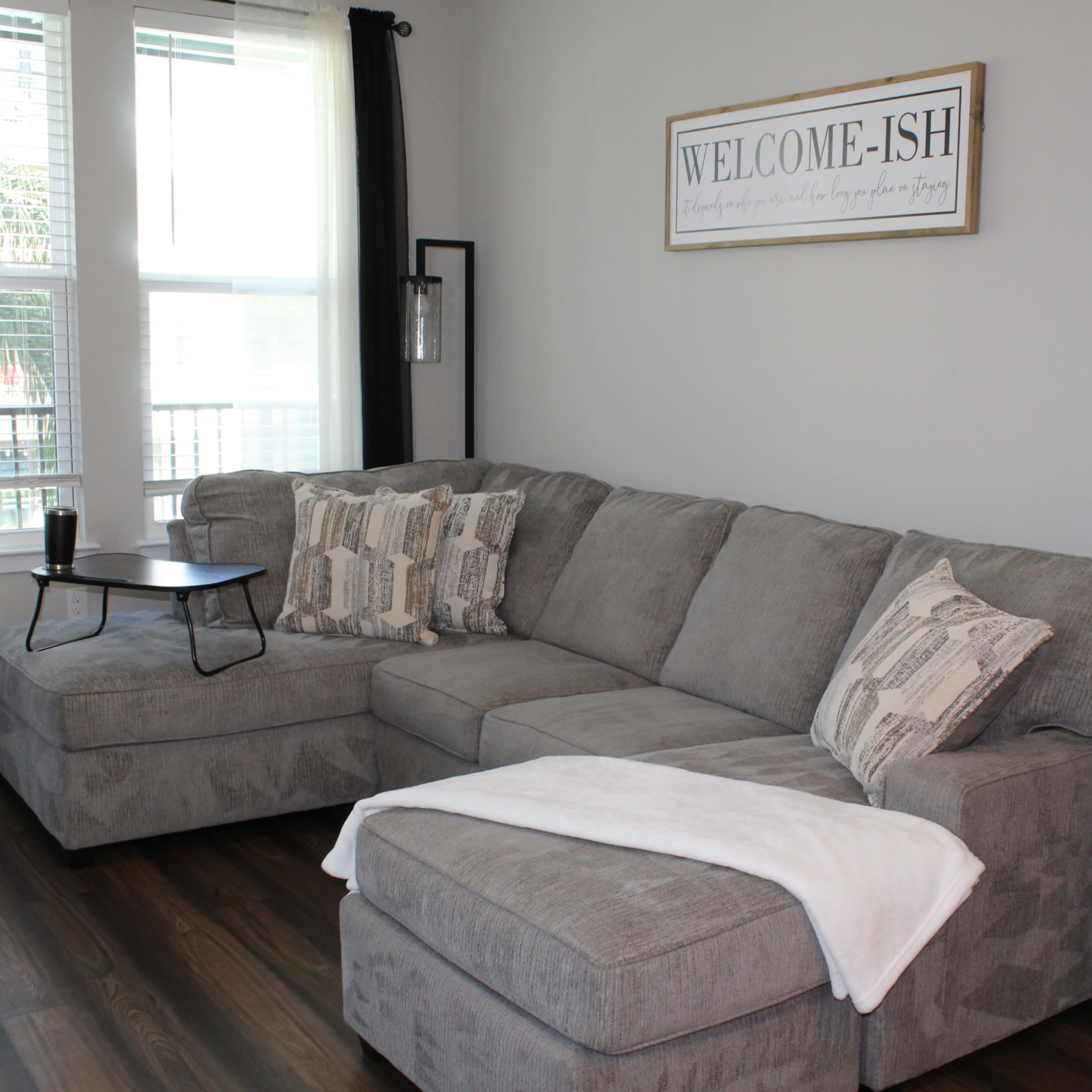 A gray sectional sofa with patterned pillows, a small side table, and a wall sign that reads "WELCOME-ISH" in a well-lit room with large windows.