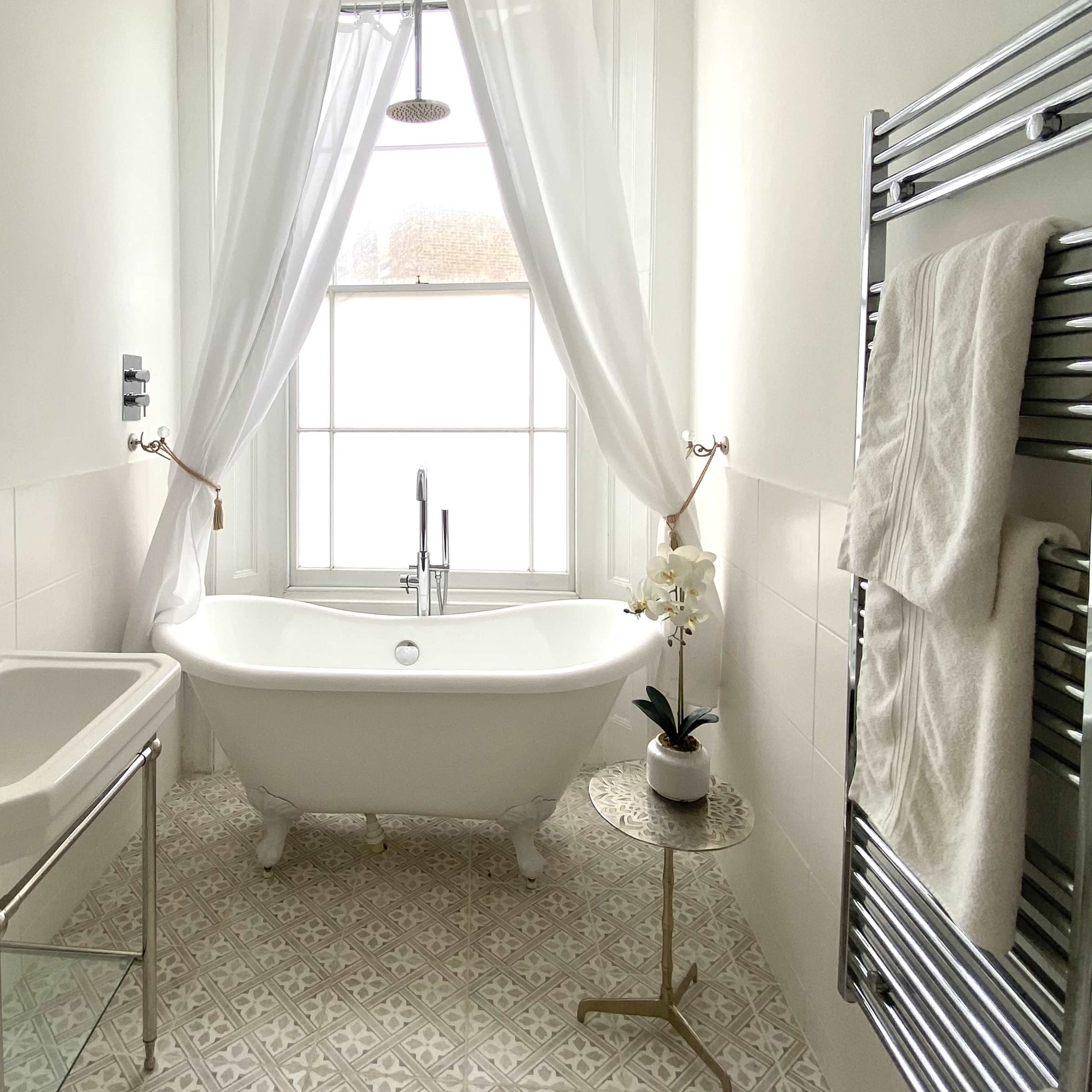 A bright, minimalist bathroom featuring a freestanding bathtub, a wall-mounted towel rack, a small side table with a potted plant, and a large window with sheer curtains.