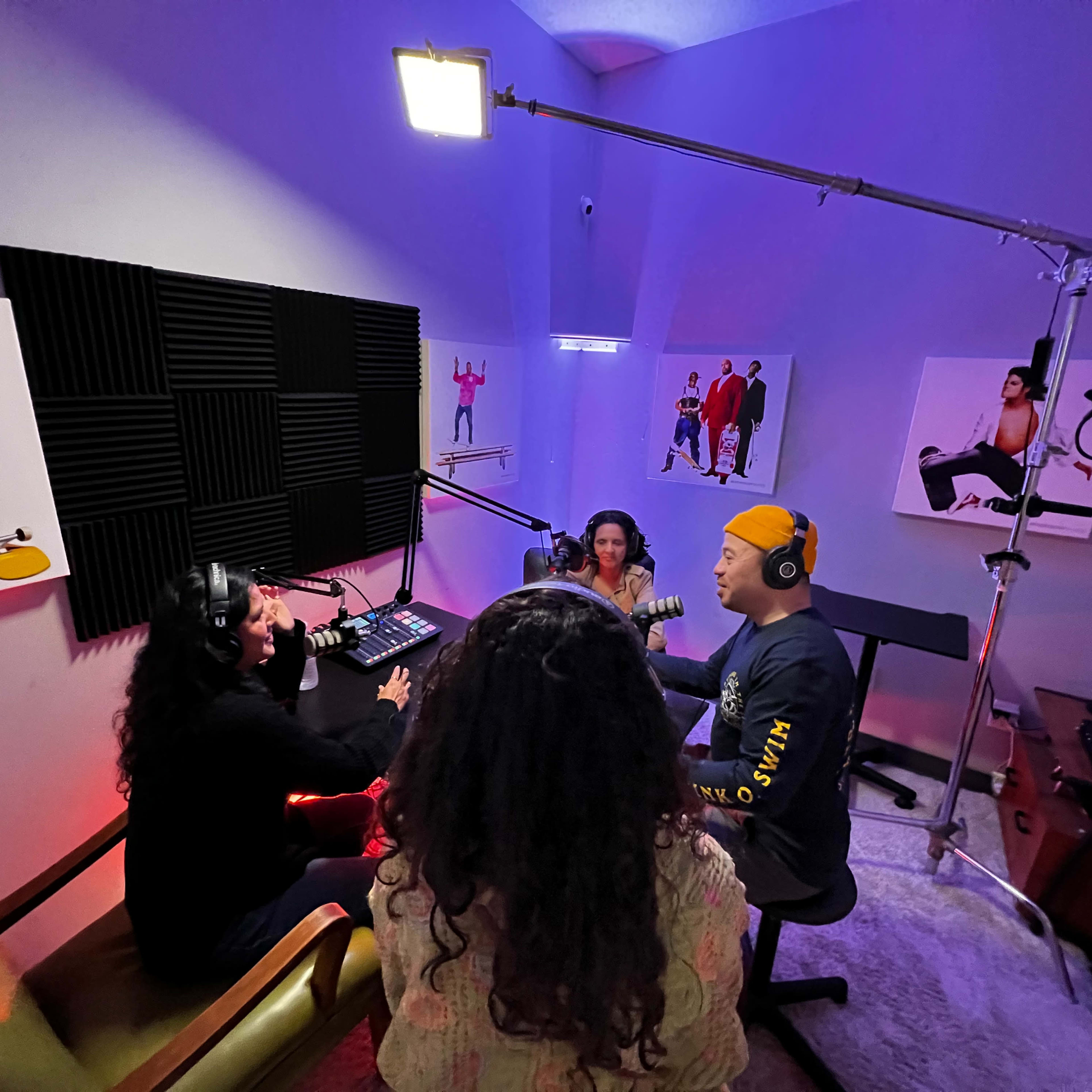 A group of three individuals is recording a podcast in a soundproofed studio with colorful lighting and artwork on the walls.