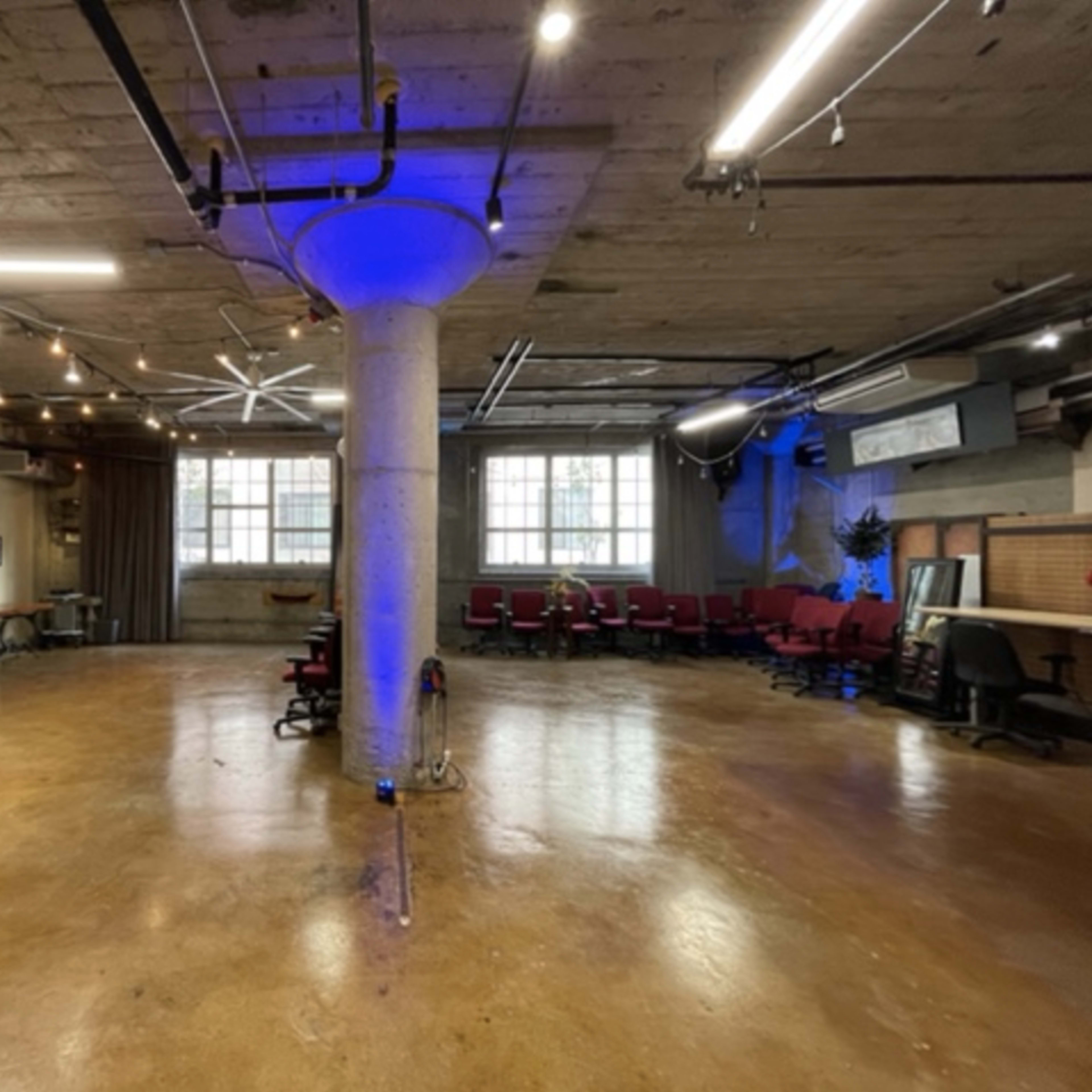 An industrial-style workspace featuring a central concrete pillar, string lights overhead, and a mix of red chairs and desks against the walls.