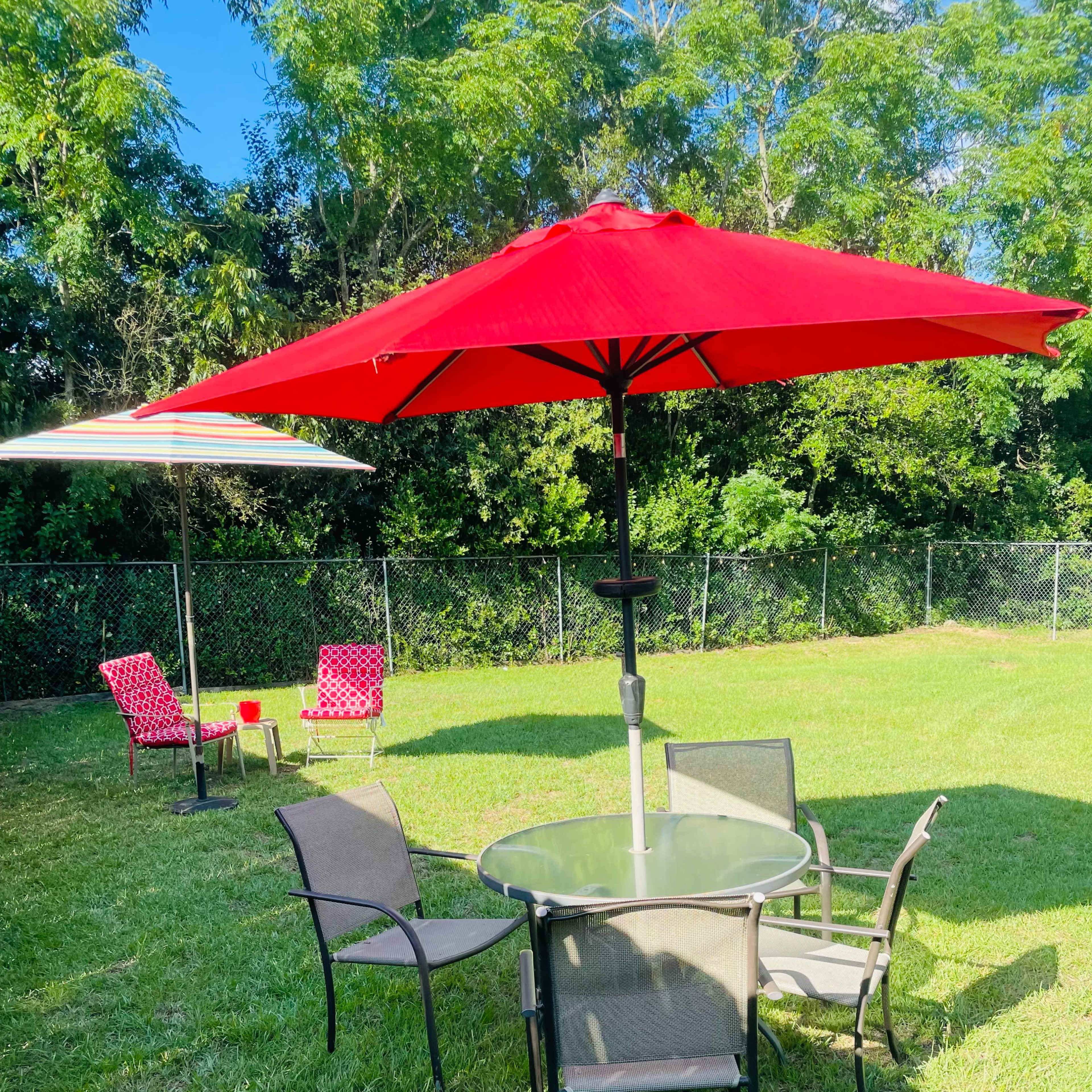 A circular glass table surrounded by six chairs is situated under red umbrellas on a grassy lawn.