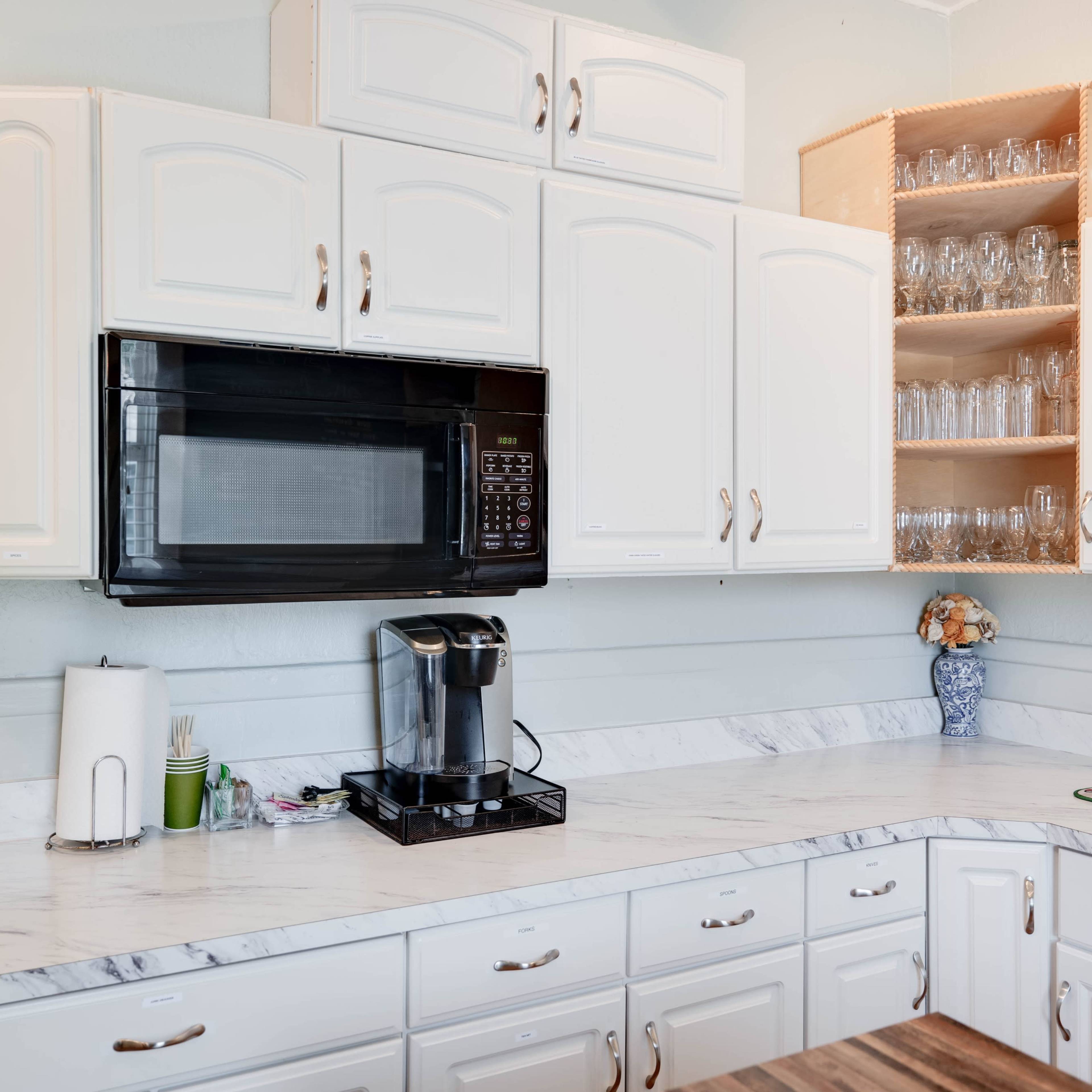 A kitchen with white cabinetry, a microwave, a coffee maker, and glassware neatly arranged on shelves above a marble countertop.