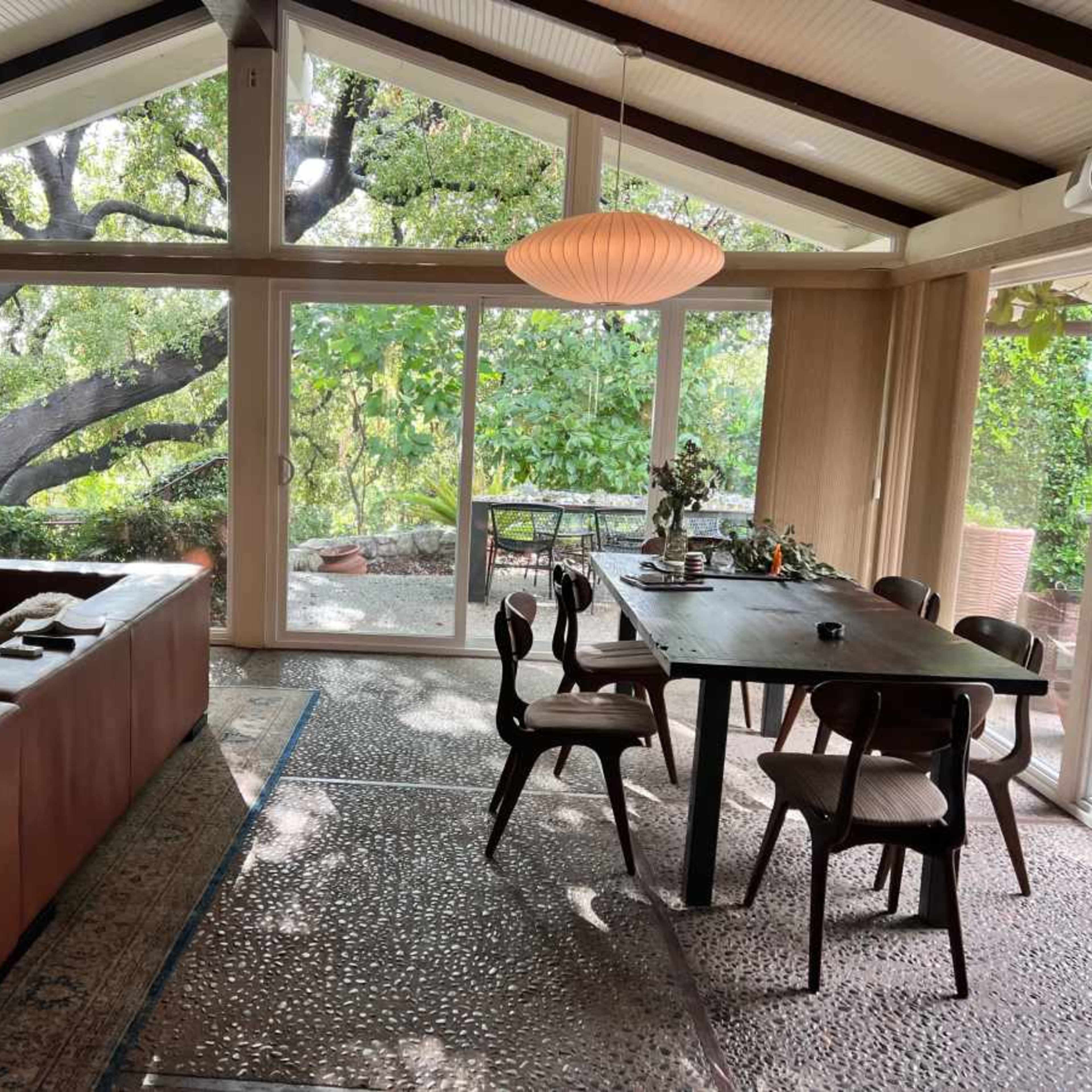 A spacious, modern living area features a glass wall with views of greenery, a wooden dining table, and a brown sofa.