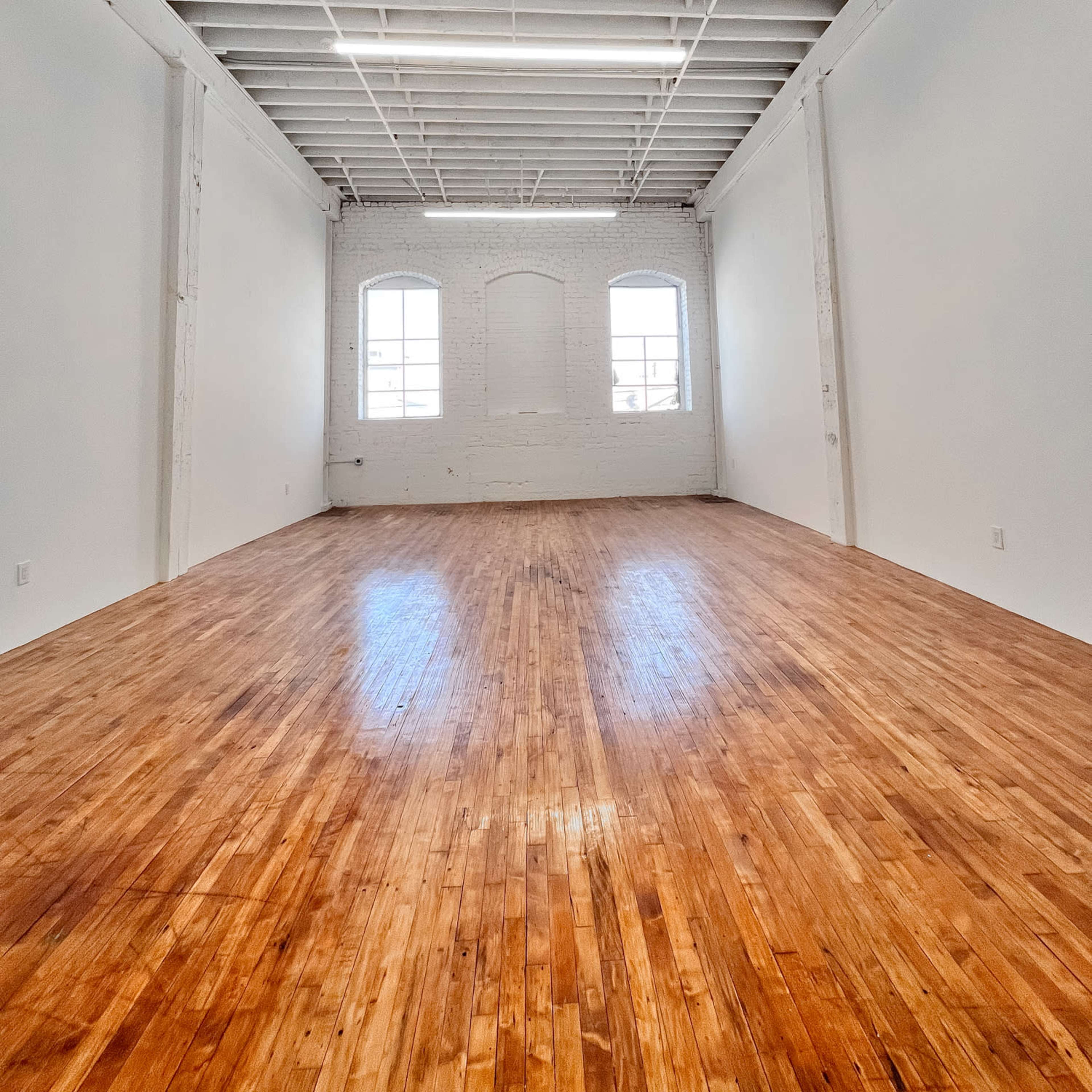 A spacious, empty room with hardwood floors and white walls, featuring large windows at one end.