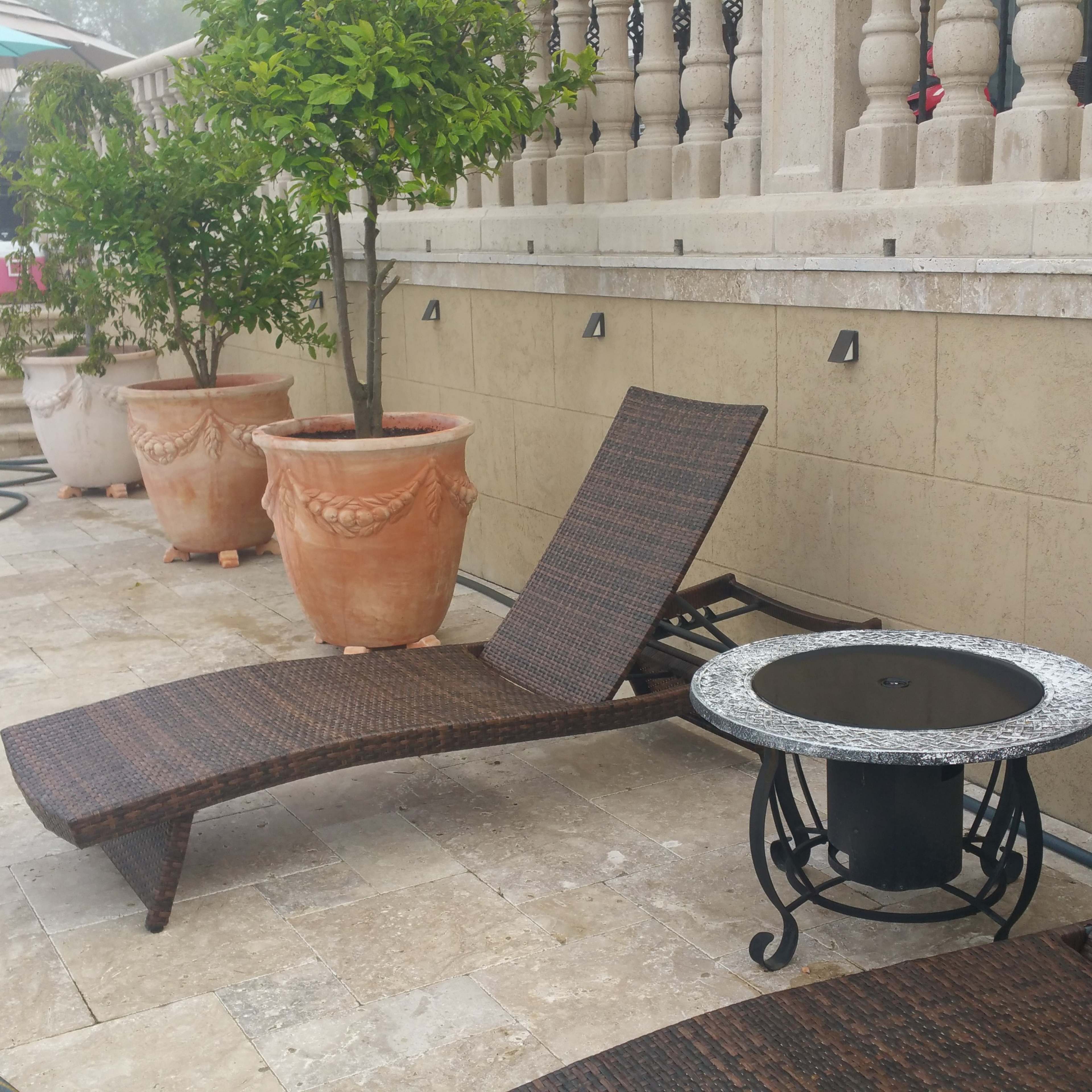 A stone patio with two lounge chairs, a small round table, and several large potted plants near a staircase.
