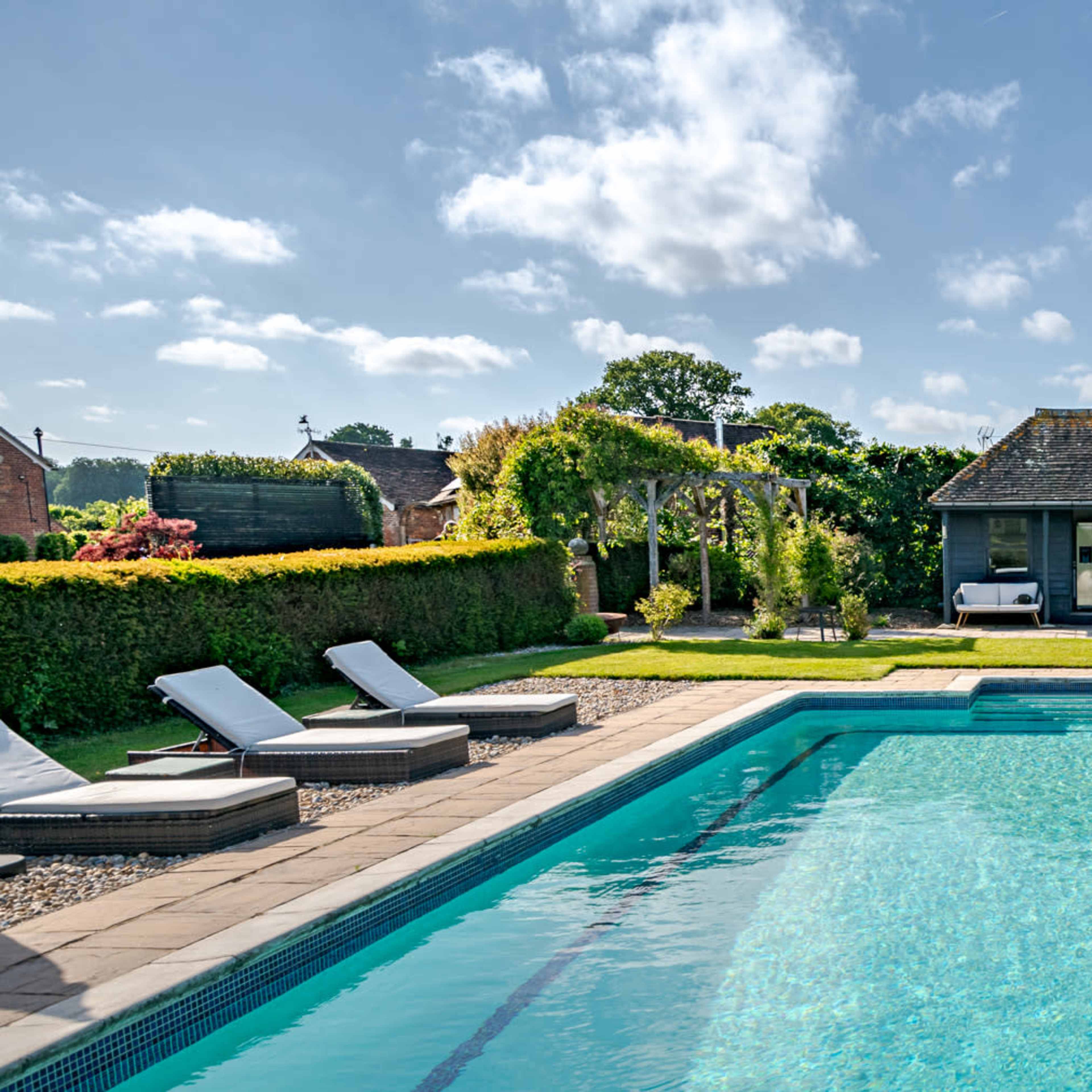 A swimming pool is surrounded by lounge chairs and a garden, with a small building in the background.