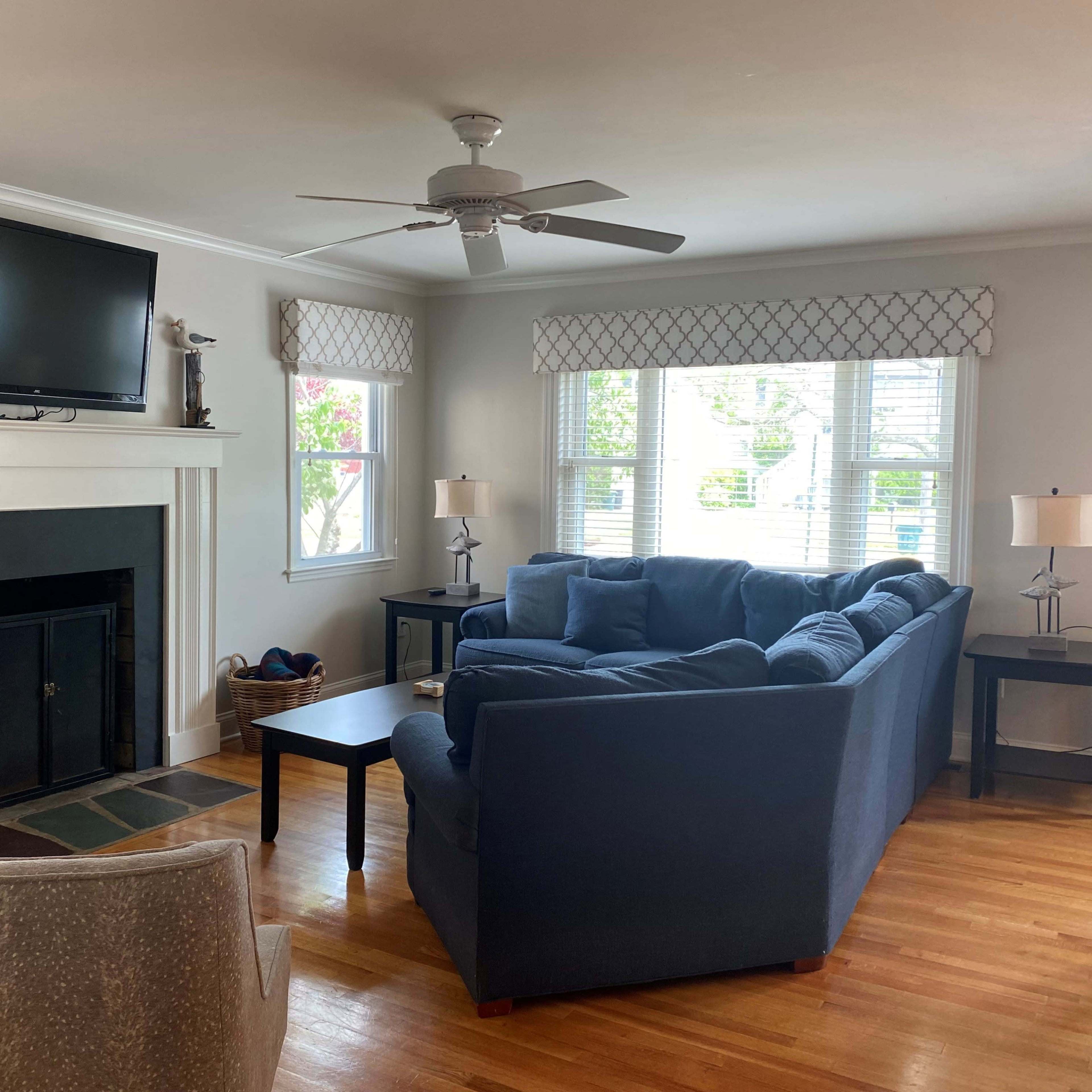 A living room features a blue sectional sofa facing a black fireplace and a television mounted above it, with windows dressed in patterned shades and two side tables with lamps.
