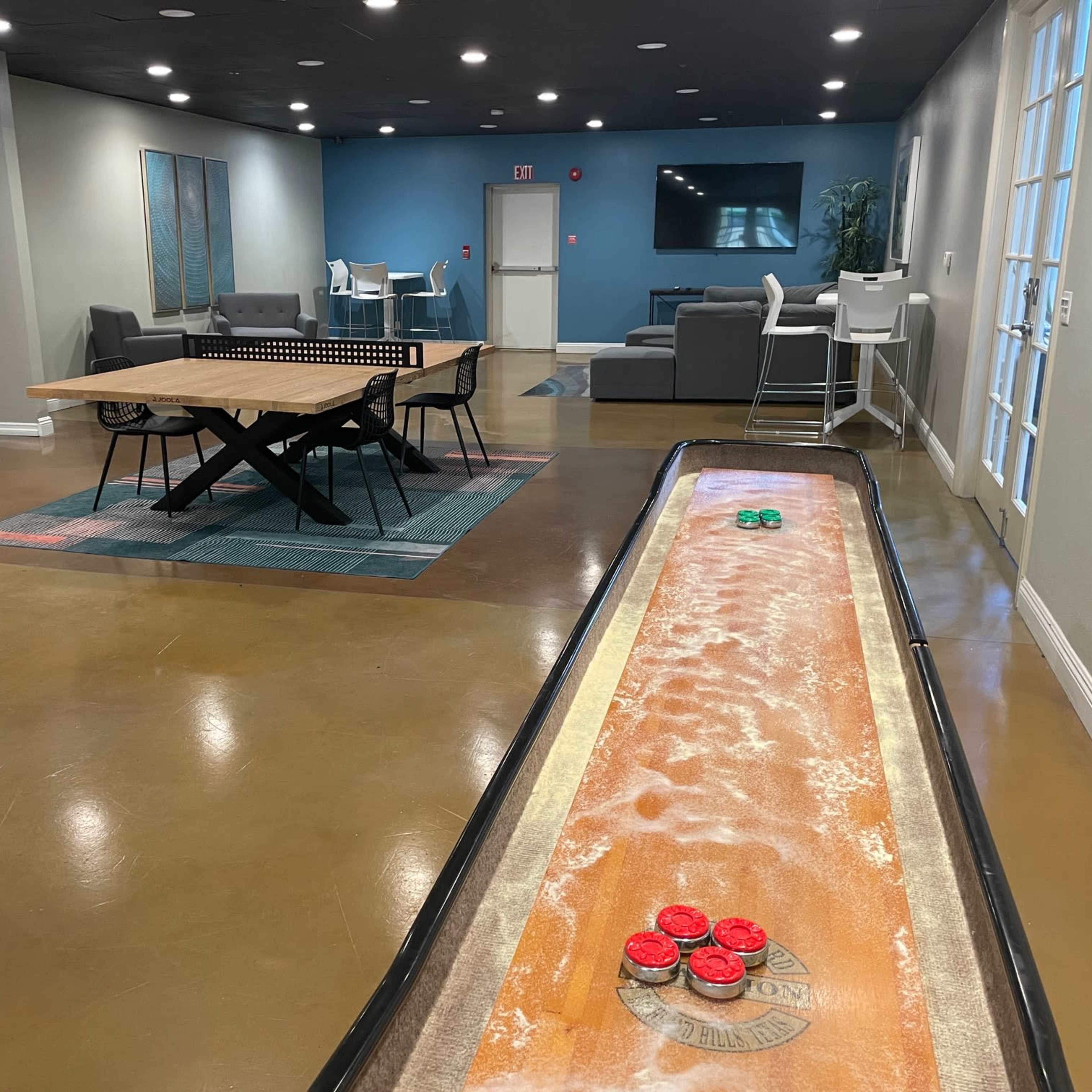 A recreational room featuring a shuffleboard table, a communal dining table, seating areas, and a large TV against the wall.
