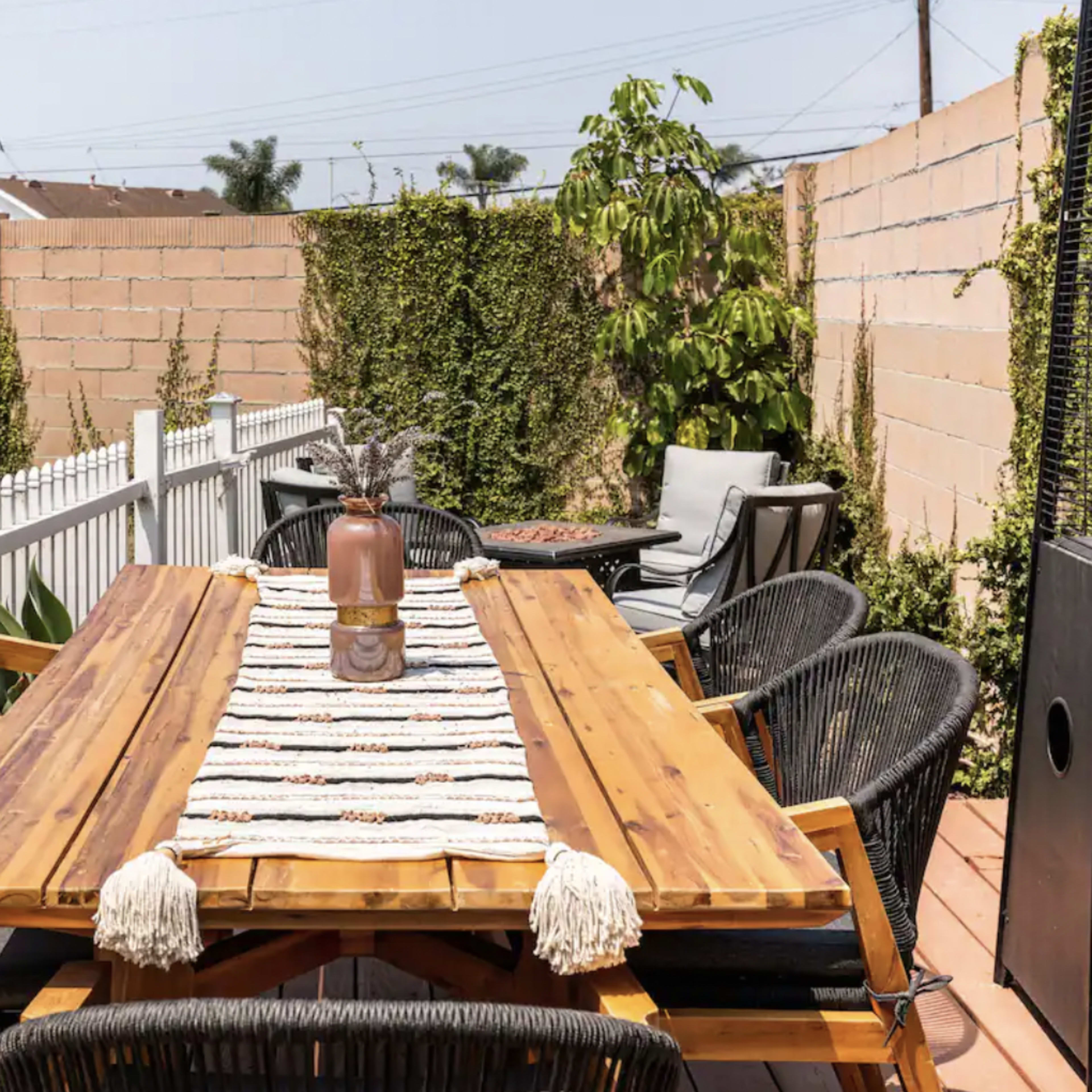 A large wooden dining table is set outdoors on a patio surrounded by black chairs and bordered by a green lawn and brick walls.