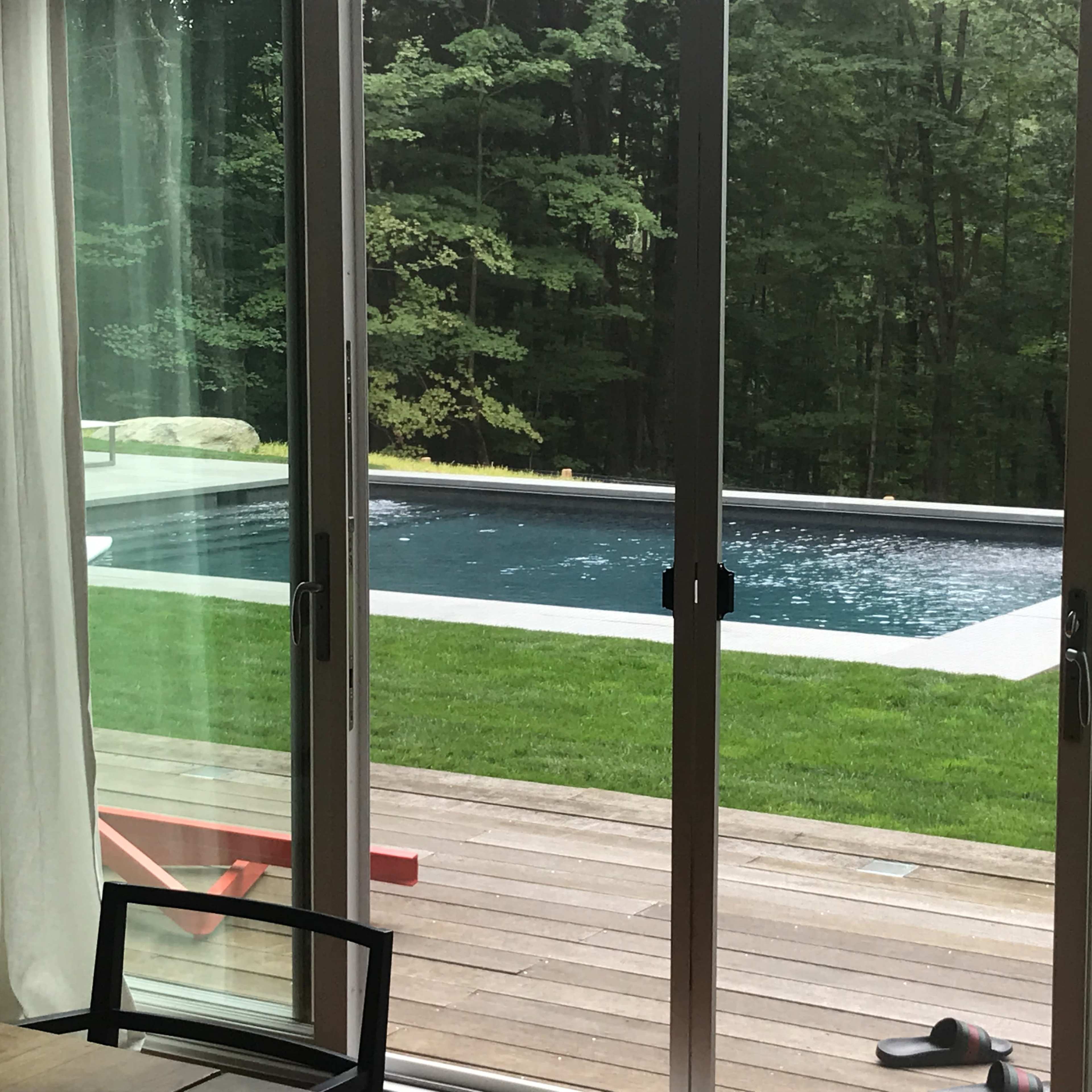 A sliding glass door frames a view of a rectangular swimming pool surrounded by grass and trees.
