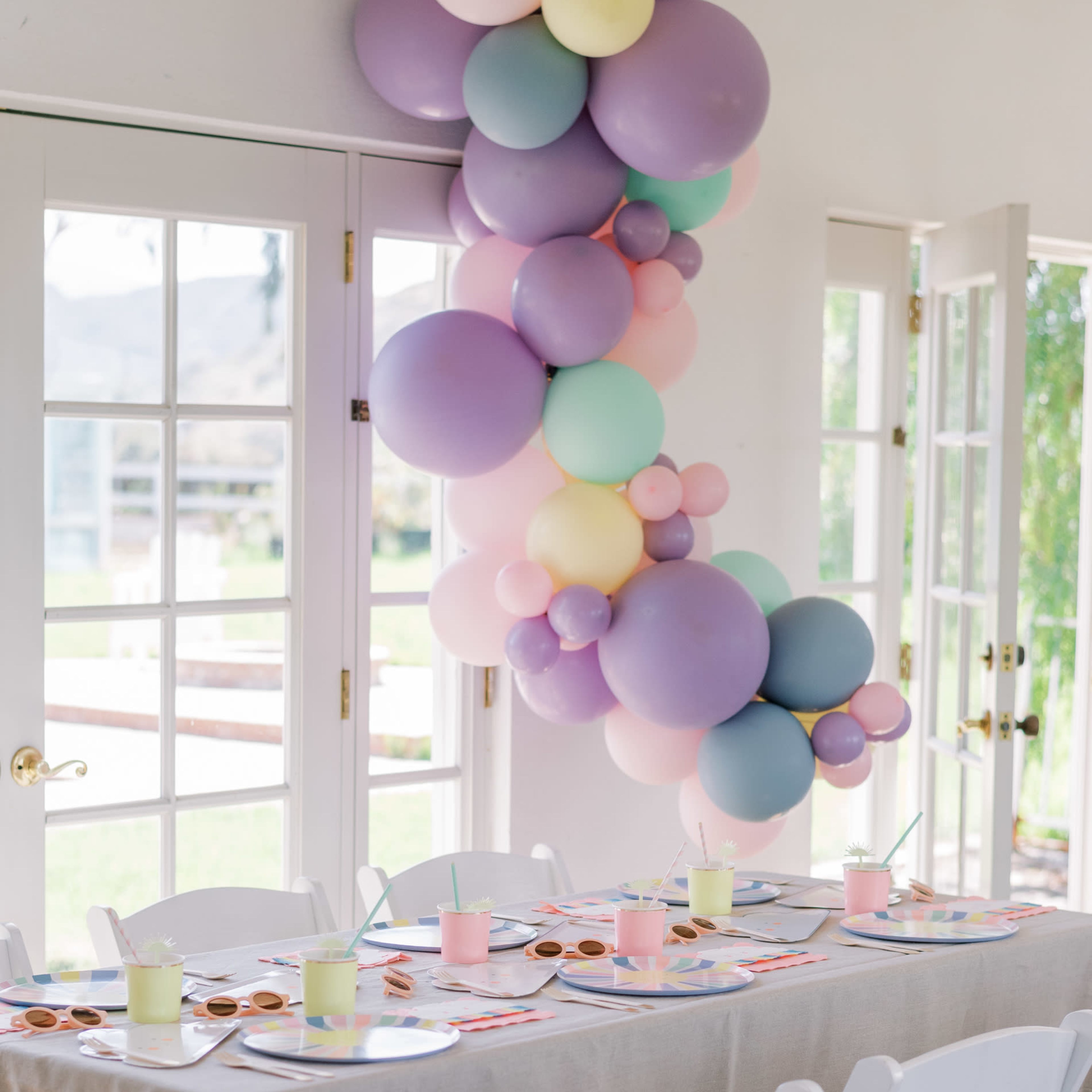 A table is set with pastel-colored plates and cups, decorated with a balloon garland, in a bright room with large windows.