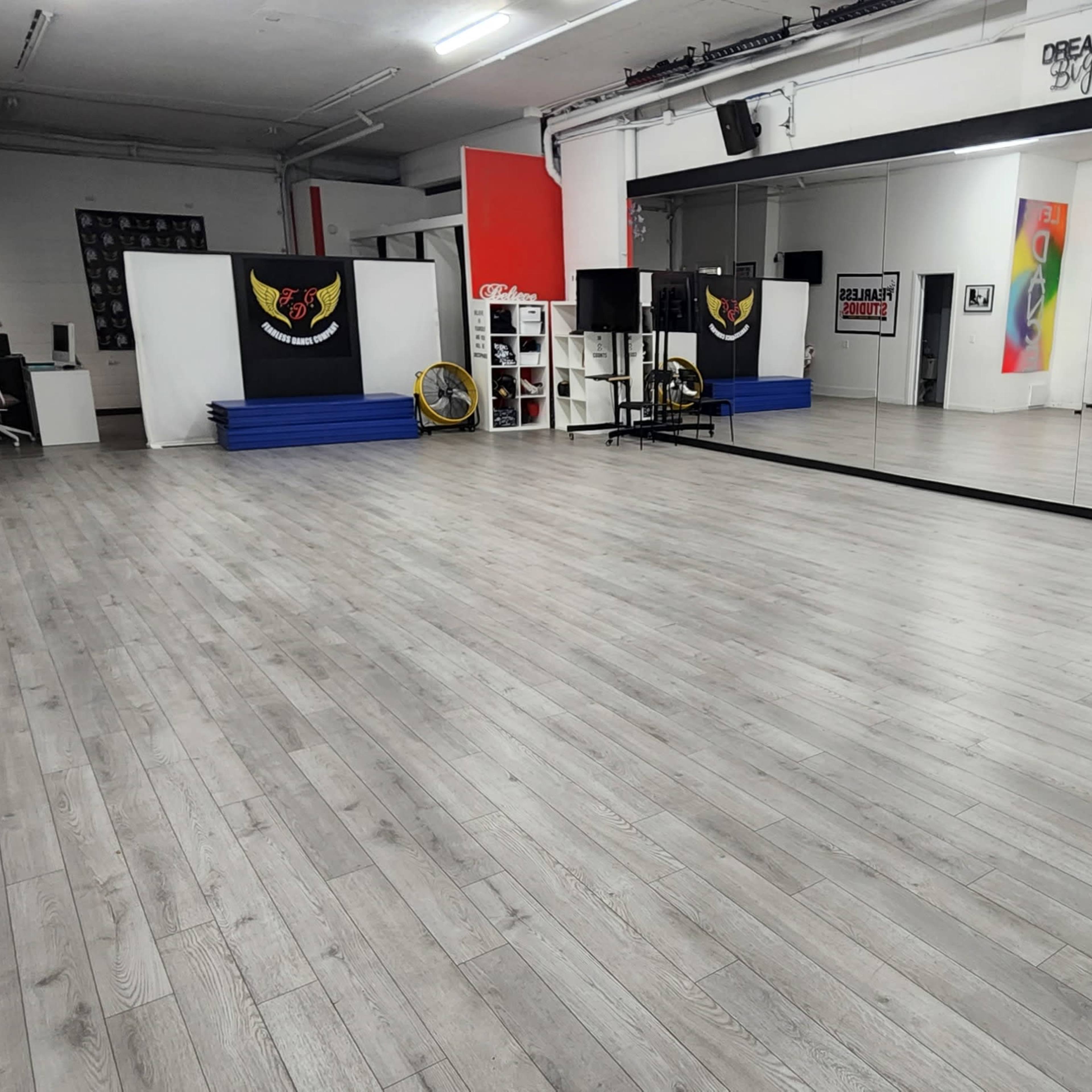 A spacious dance studio with gray wooden flooring, mirrors, and various exercise equipment arranged in a well-lit environment.